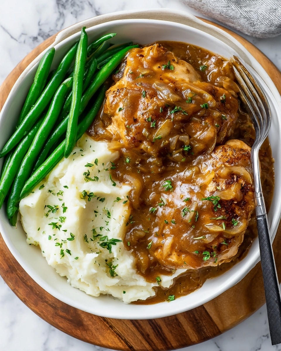 A white bowl holds a meal with three main layers: at the bottom, there is a creamy white layer of mashed potatoes on the right side; next, two pieces of browned chicken covered in thick brown onion gravy fill the center and left side, garnished with small green herb bits; at the far left side, bright green beans are arranged neatly in a row. A silver spoon with a black handle rests on the mashed potatoes on the right edge of the bowl. The bowl is placed on a round wooden board that sits on a white marbled surface. Photo taken with an iphone --ar 4:5 --v 7