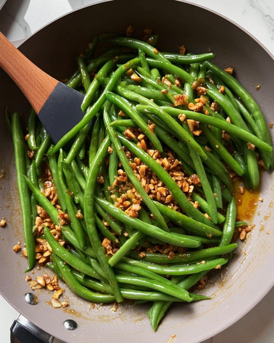 A grey frying pan holds a cooking mix of bright, fresh green beans piled on top of a base layer of finely chopped golden-brown nuts mixed with a glossy, amber sauce. The green beans are long and show a smooth texture, scattered casually over the nutty layer. A black spoon with a wooden handle is stirring from the left side, against a white marbled surface in the background. The lighting is soft and natural, highlighting the fresh and warm colors of the dish. photo taken with an iphone --ar 4:5 --v 7