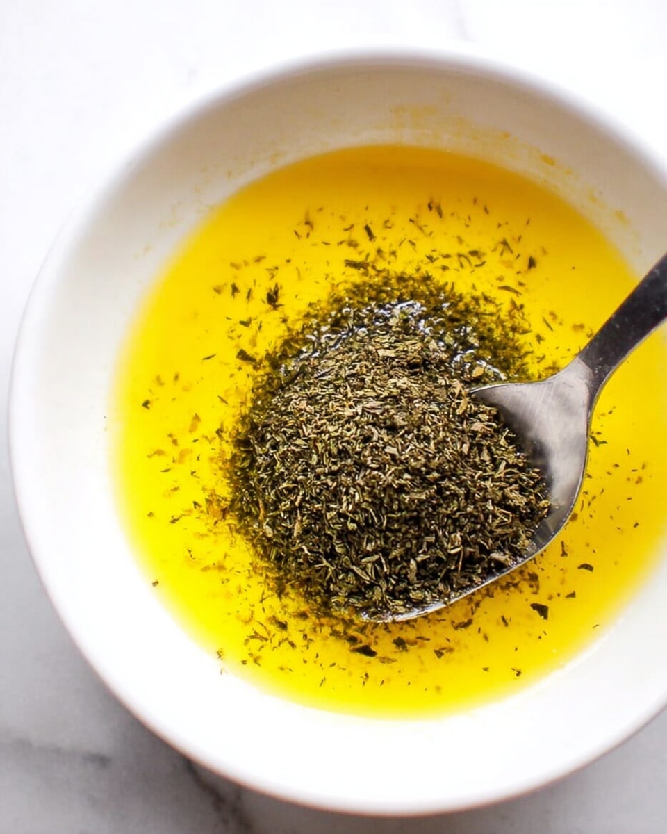 A close-up view of a white bowl contains a bright yellow liquid layer, likely melted butter or oil, covering the bottom. On top of this yellow layer, near the center, sits a mound of fine, dark green dried herbs. Resting in the bowl, angled towards the bottom right, is a metal spoon partially submerged in the yellow liquid and herbs. The bowl is placed against a white marbled texture background. photo taken with an iphone --ar 4:5 --v 7