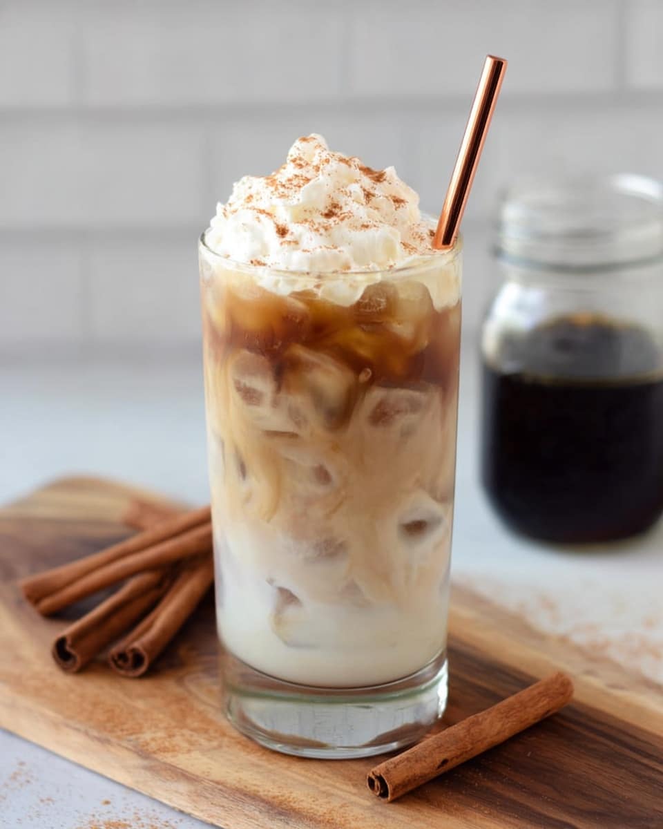 A tall clear glass shows three layers of a cold drink on a white marbled surface. The bottom layer is a dark syrup that is thin and smooth, about one inch thick. The middle layer is milky white with ice cubes floating inside, making up most of the drink. The top layer is light brown coffee swirling to meet the white milk below, filled with more ice. On top, there is a pile of whipped cream sprinkled with a light dusting of cinnamon powder. A copper-colored straw is stuck inside the whipped cream. Near the glass, three cinnamon sticks are scattered on the wooden board with a mason jar of dark syrup blurred in the background. Photo taken with an iphone --ar 4:5 --v 7