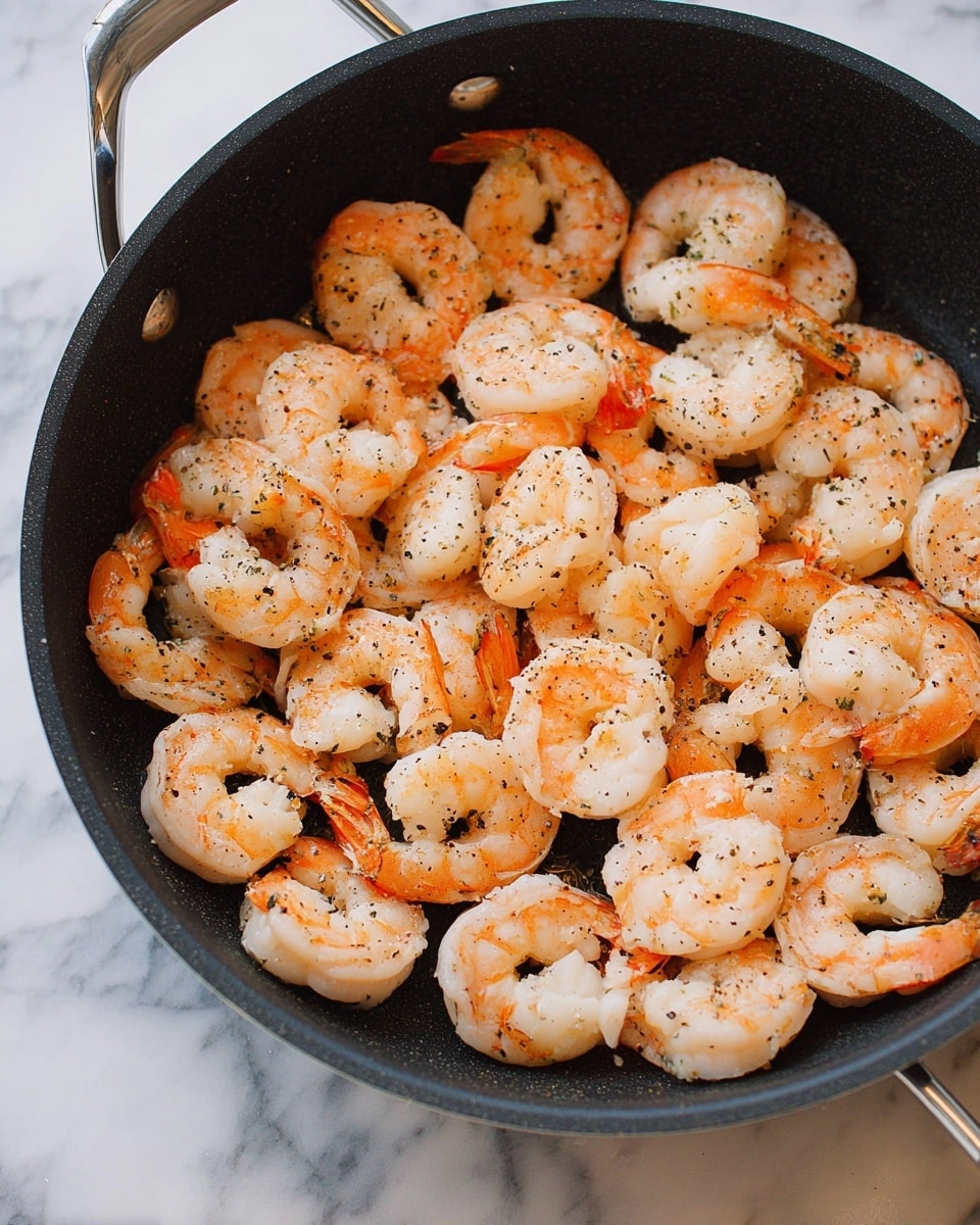 The image shows a black cooking pan filled with a single layer of cooked shrimp. The shrimp are light orange and white with visible seasoning of black pepper scattered evenly on top. Each shrimp has its tail on, curled into a loose