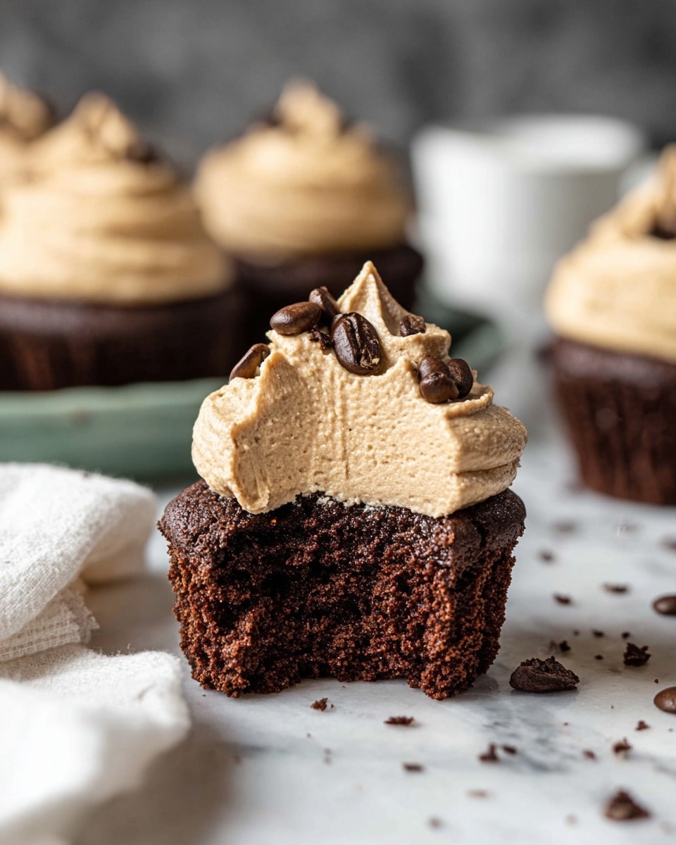 A close-up view of a chocolate cupcake with two layers: the bottom layer is a dark brown, moist textured chocolate cake, and the top layer is a thick, light brown, creamy frosting with a few coffee beans and small chocolate pieces sprinkled on it. The cupcake has one bite taken from the bottom layer, showing its soft inside. In the background, there are more cupcakes with the same frosting, blurred out and sitting on a white marbled surface. On the left side, part of a white cloth is visible, and there are some scattered coffee beans and chocolate flakes around the cupcake. Photo taken with an iphone --ar 4:5 --v 7