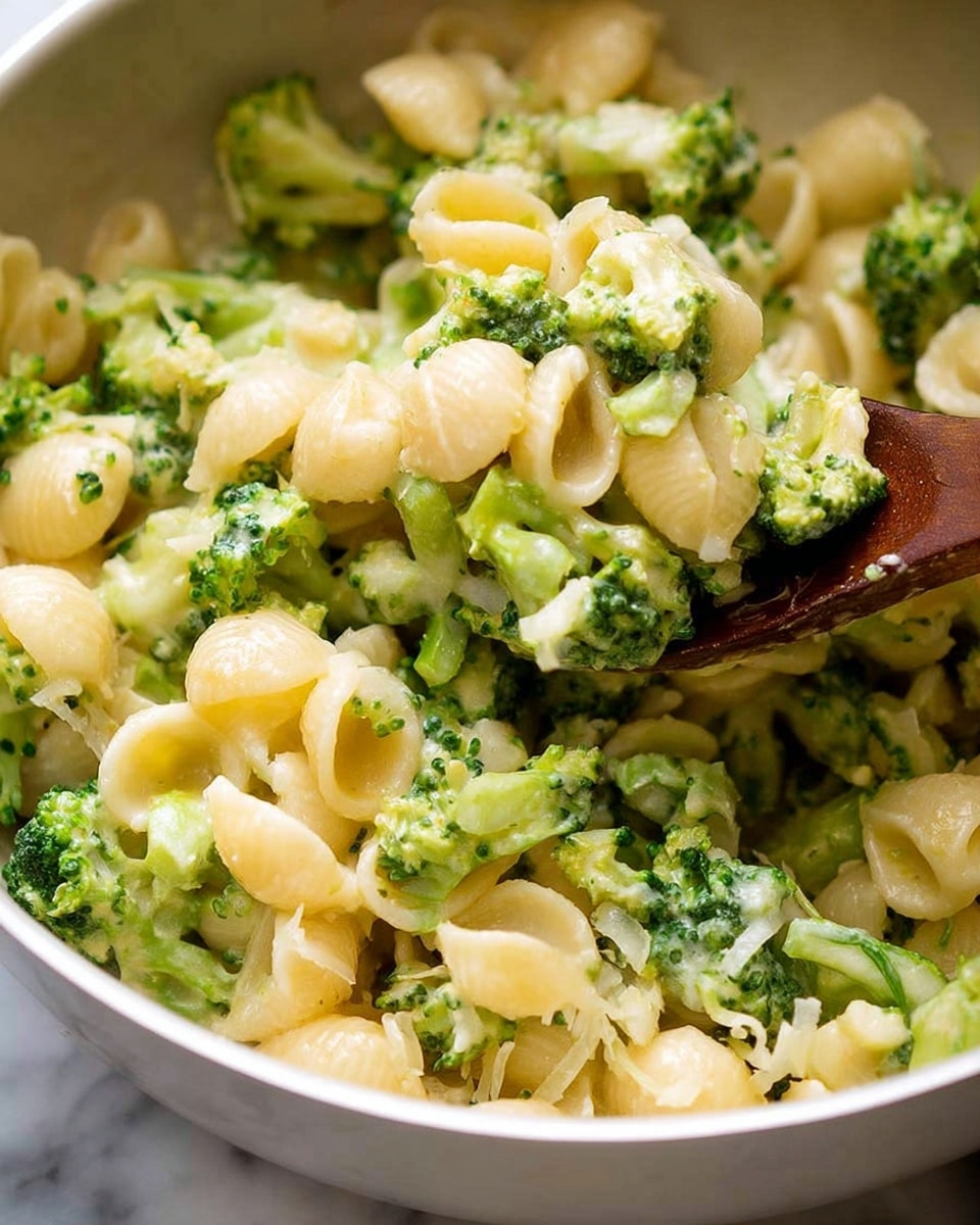 This image shows a close-up of a creamy pasta dish with green broccoli mixed evenly throughout. The pasta pieces are small, round shell shapes, pale yellow in color with a smooth and shiny texture. The broccoli is bright green, with both small chopped florets and slightly larger stalk pieces visible, giving a fresh, crunchy look. The dish looks creamy and moist, with a soft sauce coating the pasta and broccoli, creating a rich texture. A wooden spoon is partially visible, lifting some of the pasta and broccoli, adding a natural touch. The bowl holding the dish is white, and the background surface has a white marbled texture. photo taken with an iphone --ar 4:5 --v 7