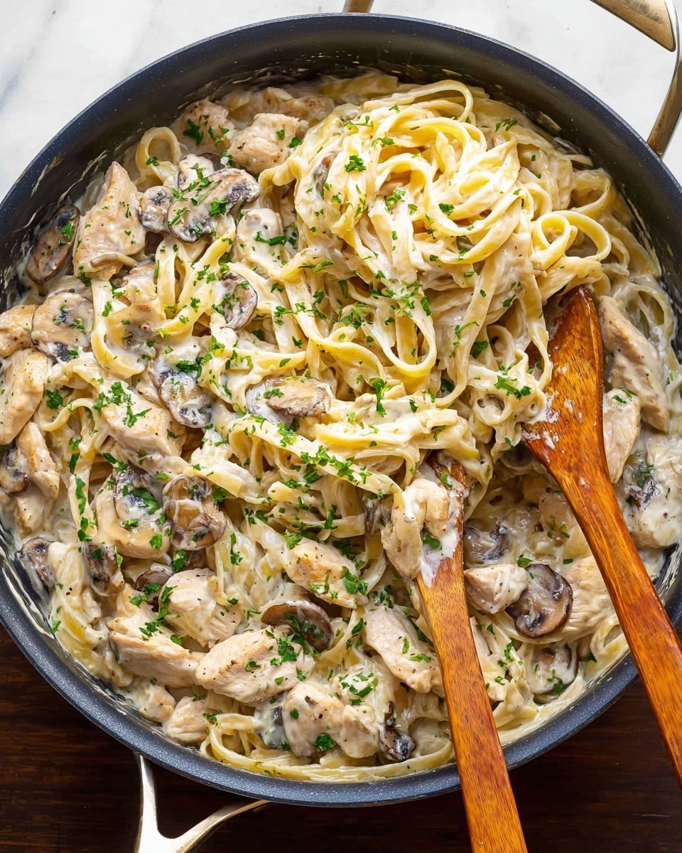 A black pan filled with creamy white sauce pasta made of long flat noodles twisted and mixed evenly with soft pieces of light beige chicken and sliced cooked mushrooms in shades of brown and gray. Two wooden spoons are placed inside the pan, lifting the pasta and chicken with some finely chopped green parsley sprinkled over the whole dish for color. The pan rests on a white marbled surface. photo taken with an iphone --ar 4:5 --v 7