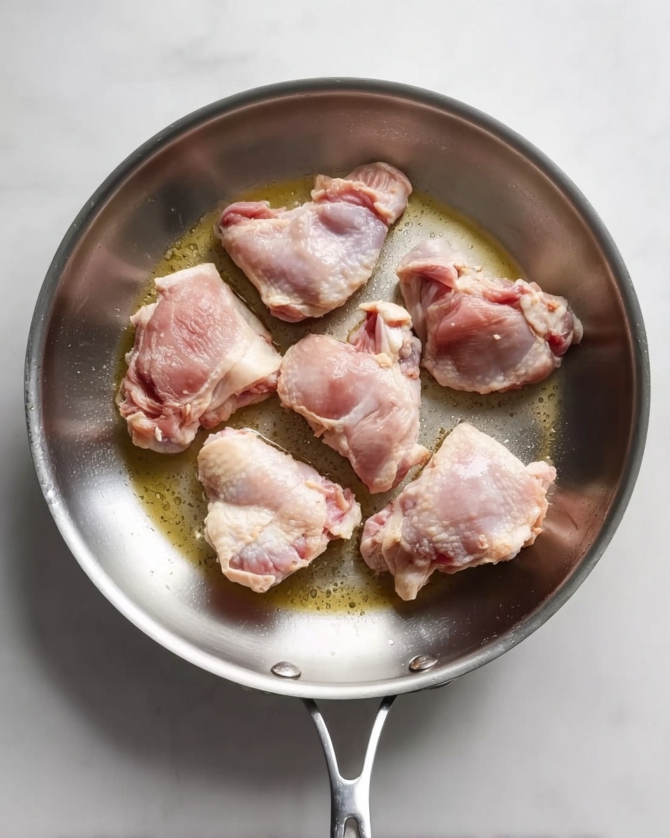 A shiny metal frying pan with six pieces of raw chicken thighs inside, arranged loosely with some space between them. The chicken pieces are pale pink with white fatty parts and small bones visible, sitting in a small amount of clear oil that has a slight golden tint. The pan is placed on a white marbled surface with soft light casting gentle shadows. photo taken with an iphone --ar 4:5 --v 7