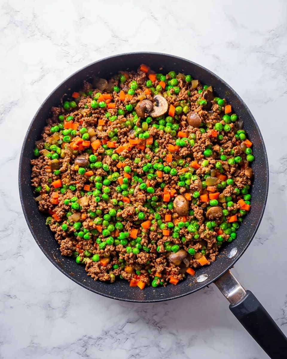 A black frying pan filled with a colorful mixture of cooked ground meat, bright green peas, small orange carrot cubes, and lightly browned small mushroom pieces, all evenly spread across the pan. The pan has a matte black handle extending out from the bottom left against a white marbled surface background. The food texture appears soft yet firm, with the green peas standing out as the brightest element on top, showing a well-mixed and cooked vegetable and meat combination. Photo taken with an iphone --ar 4:5 --v 7