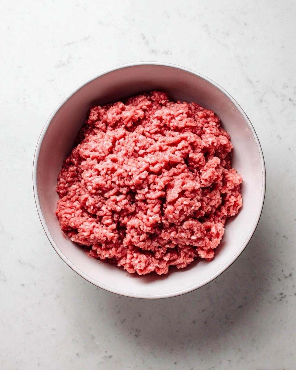A round white bowl filled with raw ground meat, showing a coarse texture with small chunks and a vibrant pinkish-red color. The bowl is placed on a white marbled surface with faint grey veins, giving a clean and bright look. The meat is evenly spread inside the bowl, creating a soft, uneven surface. No other objects are visible in the scene. photo taken with an iphone --ar 4:5 --v 7
