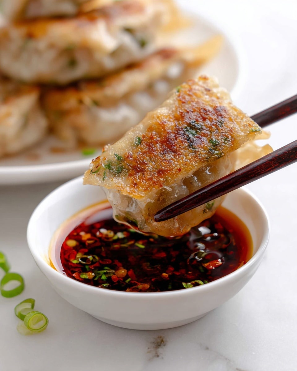 The image shows a close-up of a piece of square dumpling being held by dark brown chopsticks above a small white bowl filled with a dark red dipping sauce containing chili flakes and green onion slices. The dumpling has a golden-brown crispy surface with visible green herbs and is wrapped in a translucent skin that shows the texture of the cooked filling inside. The bowl sits on a white marbled surface with some green onion pieces scattered nearby. In the background, there is a white plate holding more dumplings stacked closely together, slightly blurred. photo taken with an iphone --ar 4:5 --v 7