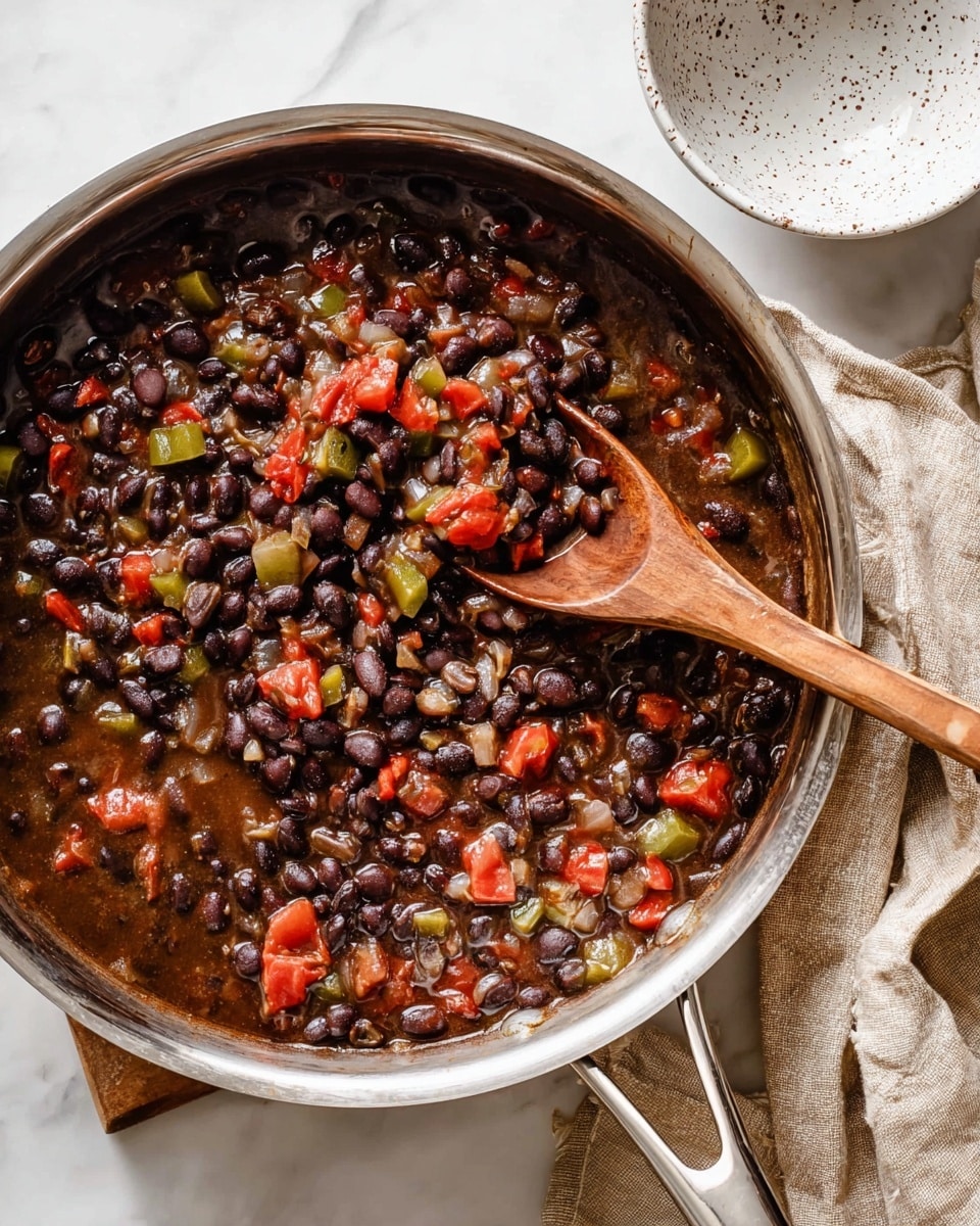 A stainless steel pan filled with a thick mix of black beans, diced green peppers, chopped onions, and chunks of red tomatoes in a rich, dark broth. A wooden spoon rests inside the pan, partly covered with the bean mixture, showing some beans and tomatoes on it. The pan is placed on a white marbled surface with a folded beige cloth beside it, and a white speckled bowl sits near the top right corner. photo taken with an iphone --ar 4:5 --v 7