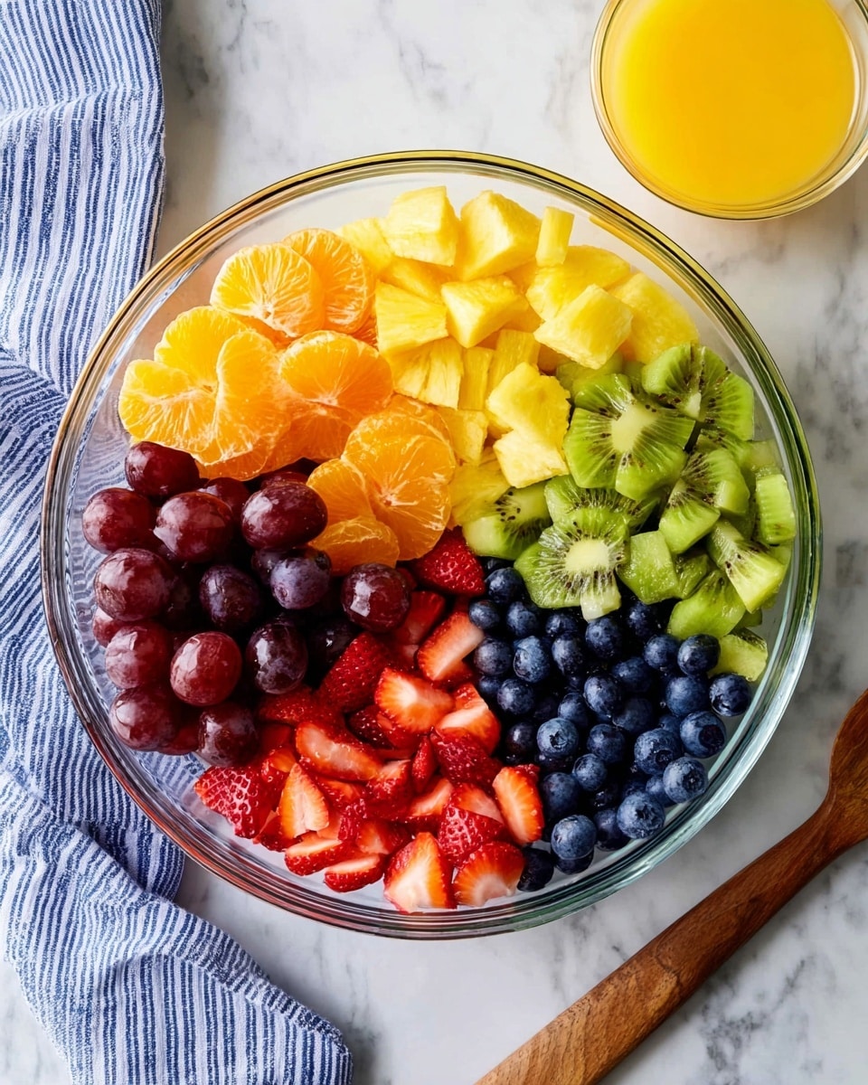 A clear glass bowl on a white marbled surface holds six sections of fresh fruit arranged in a circle: deep purple grapes at the bottom left, bright orange mandarin slices next to them, then yellow pineapple chunks to the right, green kiwi pieces at the top right, dark blue blueberries at the top left, and red sliced strawberries in the center. A small glass bowl with orange juice sits near the top right of the scene, and a blue-and-white striped cloth is partially visible on the left side. A wooden spoon handle appears at the bottom right edge. Photo taken with an iphone --ar 4:5 --v 7