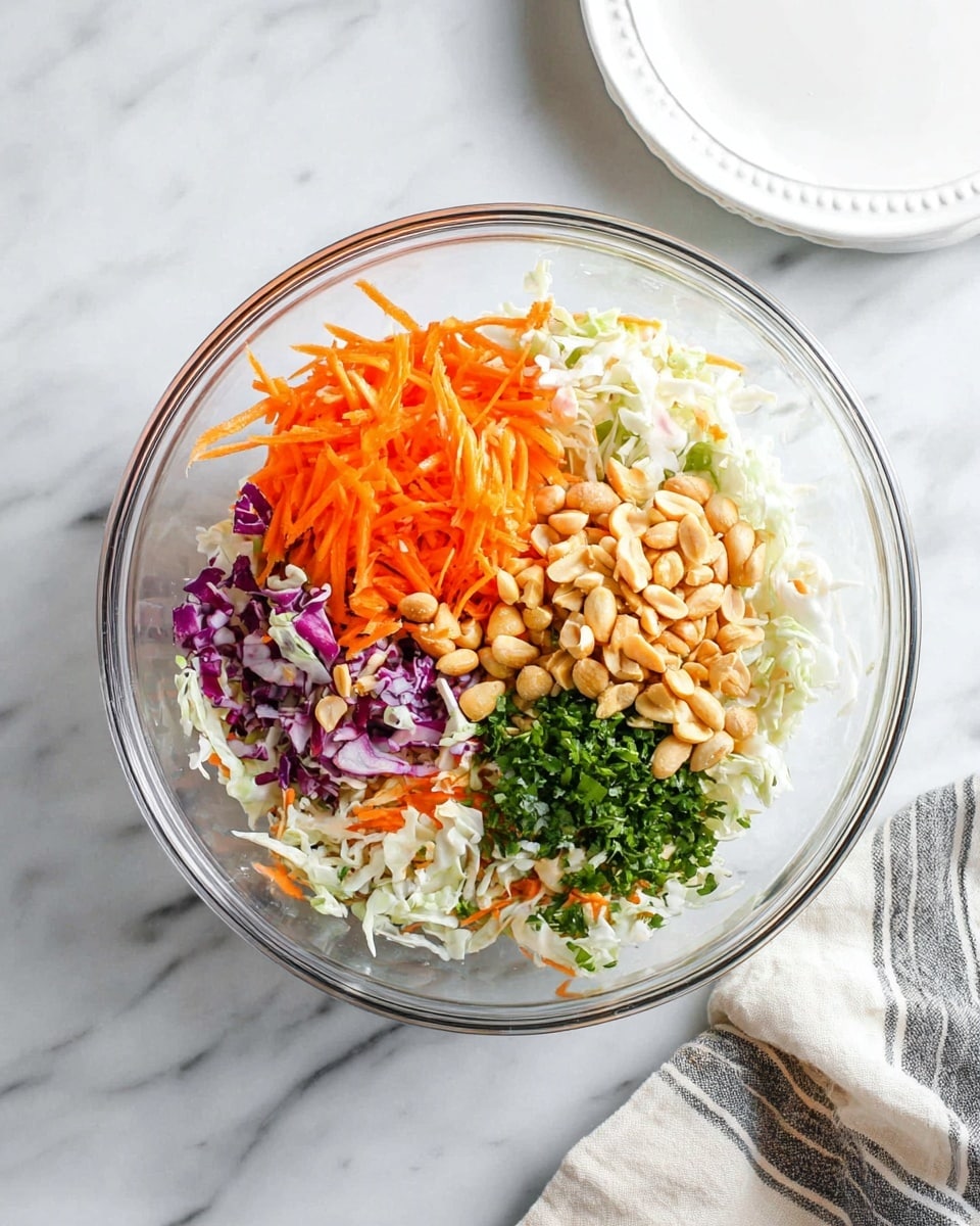 A clear glass bowl holds a fresh mixture of ingredients layered side by side. At the bottom is a base of shredded white and purple cabbage mixed with small orange carrot pieces. On top of that, there are bright orange shredded carrots stacked in one corner. Next to the carrots, there is a pile of light brown peanuts. Beside the peanuts, finely chopped green herbs add a vibrant touch. The bowl sits on a white marbled surface with a white plate and a striped cloth napkin nearby. photo taken with an iphone --ar 4:5 --v 7