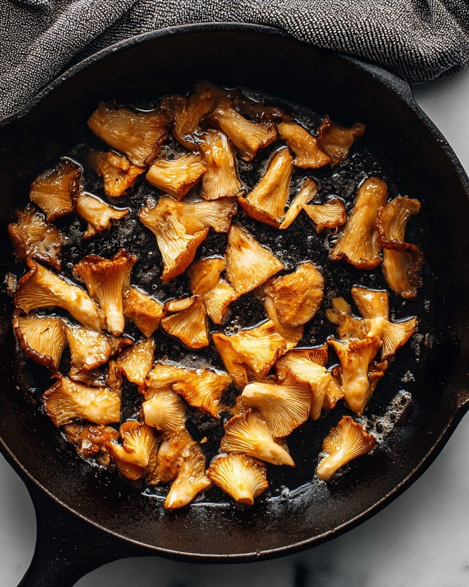 The image shows golden-brown oyster mushrooms cooking in a black cast-iron skillet. The mushrooms are cut into small, uneven pieces with a mix of light beige and deeper brown colors, slightly crispy and glossy from the cooking oil. The skillet holds around 20 pieces spread out evenly, with small bubbles of oil around them. The background is a white marbled surface with a gray textured cloth partially visible on the top left corner. photo taken with an iphone --ar 4:5 --v 7