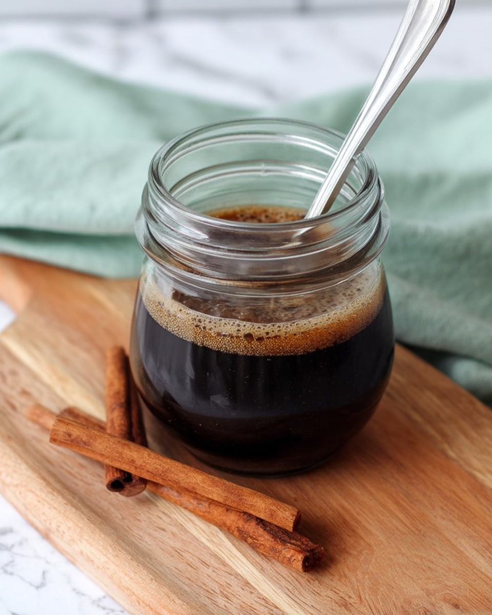 A small clear glass jar filled about two-thirds with dark brown syrupy liquid with a slight frothy layer on top, placed on a light wooden board with two whole cinnamon sticks lying beside it; a silver spoon rests inside the jar. The background has a white marbled texture surface and a light green cloth is softly blurred behind the jar. Photo taken with an iphone --ar 4:5 --v 7