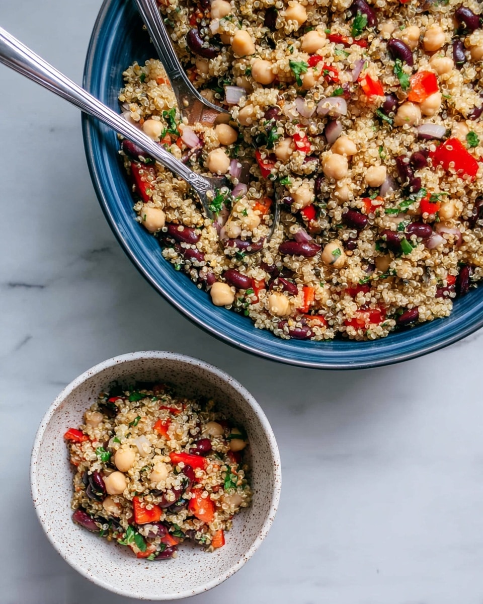 The image shows a large blue bowl filled with a colorful quinoa salad made from light beige quinoa grains mixed with three types of beans: pale beige chickpeas, small black beans, and white beans. The quinoa and beans are mixed with bright red bell pepper pieces, small green herb leaves, and small pieces of white onion, creating a speckled and textured look. Two shiny silver spoons rest inside the large bowl, one of which reaches into a smaller white speckled bowl below that is also filled with the same quinoa salad. The scene is set on a white marbled surface, highlighting the colors and textures of the quinoa salad. Photo taken with an iphone --ar 4:5 --v 7