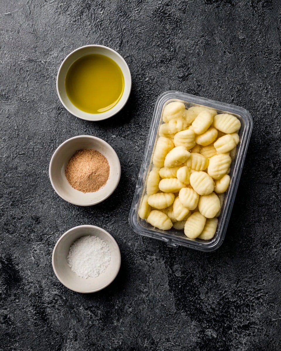 The image shows a clear plastic package filled with pale yellow gnocchi placed on a dark textured surface. To the left of the package are three small white bowls arranged vertically: the top bowl contains a light golden yellow liquid that looks like olive oil, the middle bowl holds a light brown powder resembling yeast, and the bottom bowl has white granules that appear to be salt. The overall scene is minimal with focus on the ingredients. photo taken with an iphone --ar 4:5 --v 7