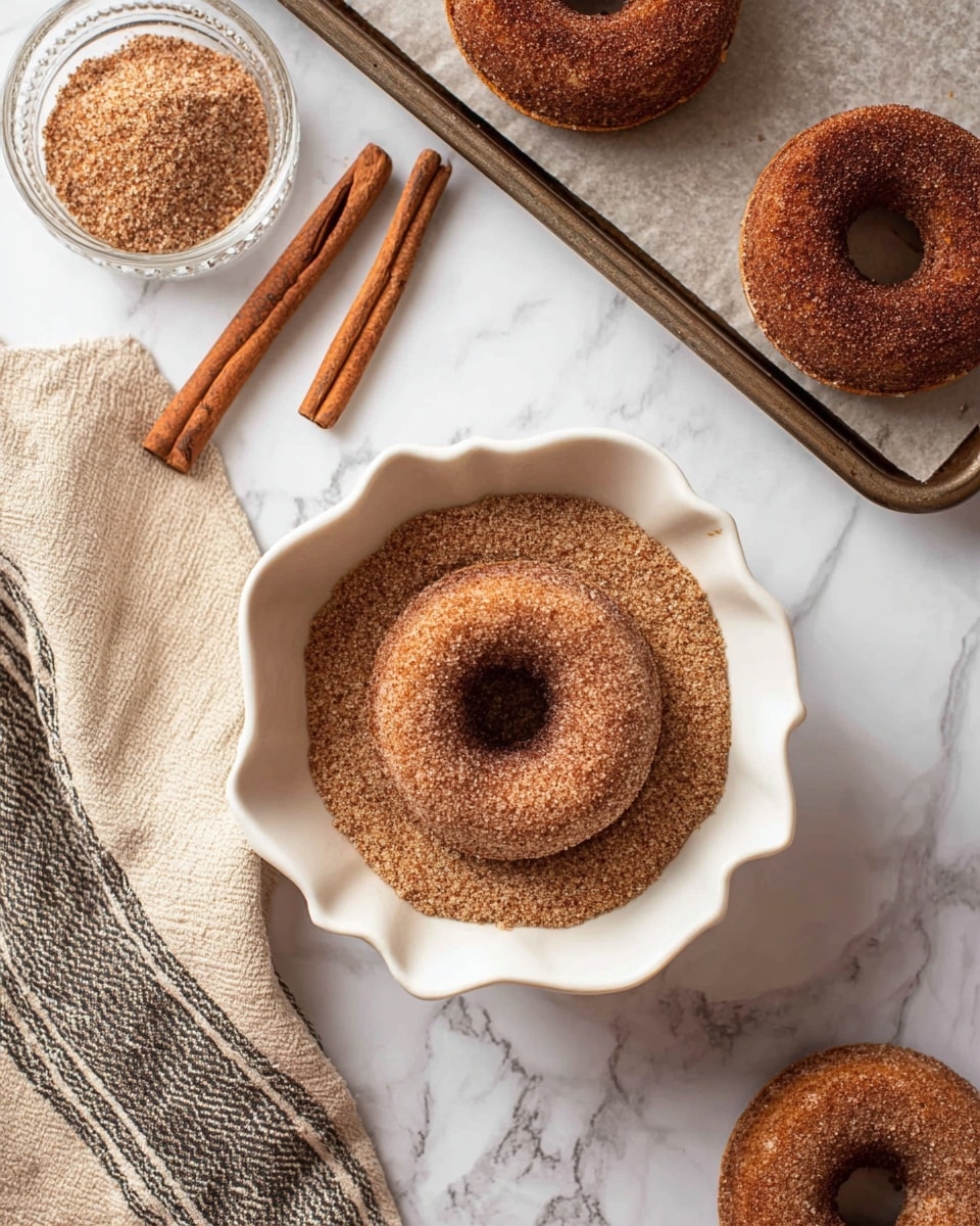 The image shows a cinnamon sugar coated donut sitting in a white scalloped bowl filled with a layer of cinnamon sugar mix. Around the bowl, there are two whole cinnamon sticks on a white marbled surface. To the right, two more cinnamon sugar donuts rest on a parchment-lined baking tray with a metal edge. Below the bowl, there is another plain donut placed on the white marbled surface next to a beige cloth with dark stripes. The overall color scheme is warm brown tones of the donuts and cinnamon sticks, contrasted by the white bowl and white marbled surface. Photo taken with an iphone --ar 4:5 --v 7
