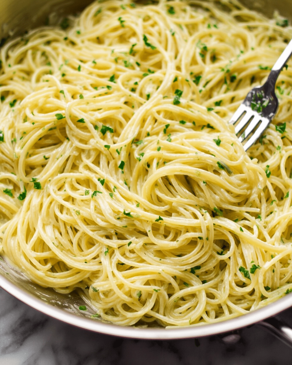 A close-up view of a single layer of cooked spaghetti pasta mixed with light creamy sauce, scattered with finely chopped green herbs, all inside a shallow pan. The pasta strands are twisted and intertwined, showing a smooth, slightly glossy texture. A silver fork is partially visible resting among the noodles, adding a bit of metallic shine. The background is a soft white marbled surface. photo taken with an iphone --ar 4:5 --v 7