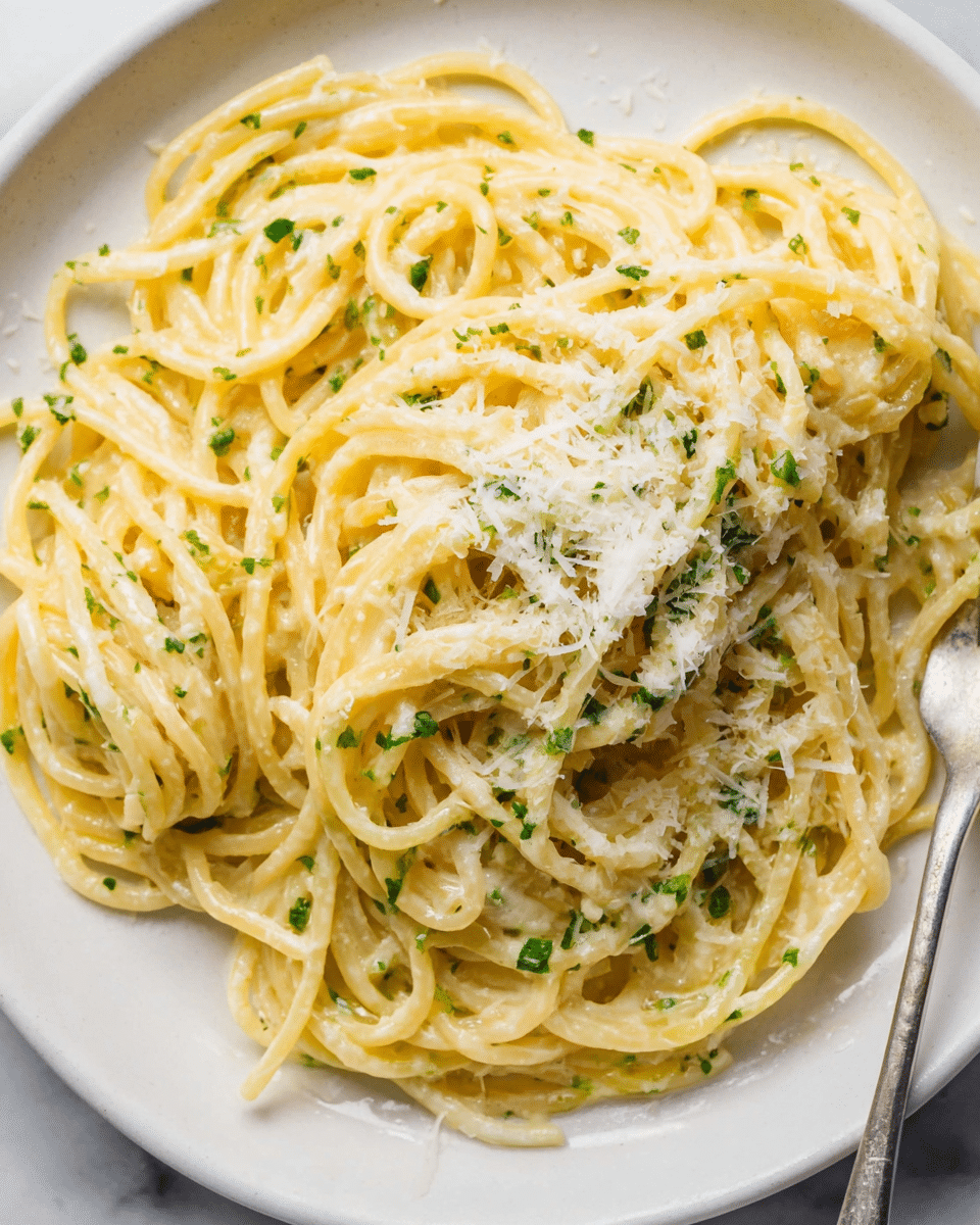 A close-up view of a white plate filled with a single large serving of creamy spaghetti. The pasta is light yellow, coated in a smooth creamy sauce, with small bits of green herbs mixed evenly throughout. On top, there is a generous light layer of finely shaved white cheese, adding texture contrast. The plate rests on a white marbled surface and a silver fork is partially visible at the plate's edge. Photo taken with an iphone --ar 4:5 --v 7