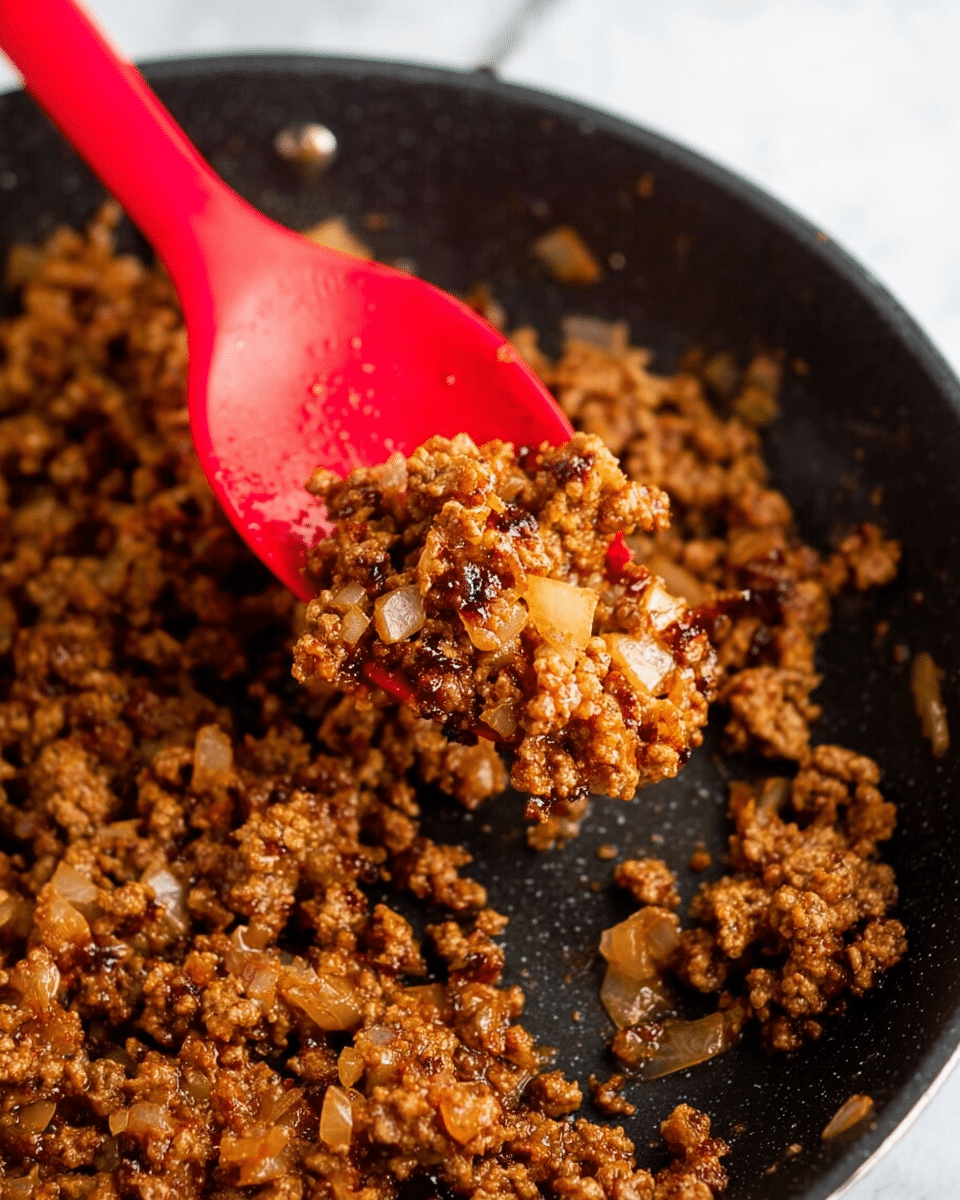 A close-up view of cooked ground meat mixed with small pieces of translucent, browned onions in a black pan. The mixture is coarse and crumbly with a rich brown color, showing some slight caramelization. A bright red silicone spoon is lifting some of the mixture, adding a pop of color to the dark pan. The background is a white marbled texture. photo taken with an iphone --ar 4:5 --v 7