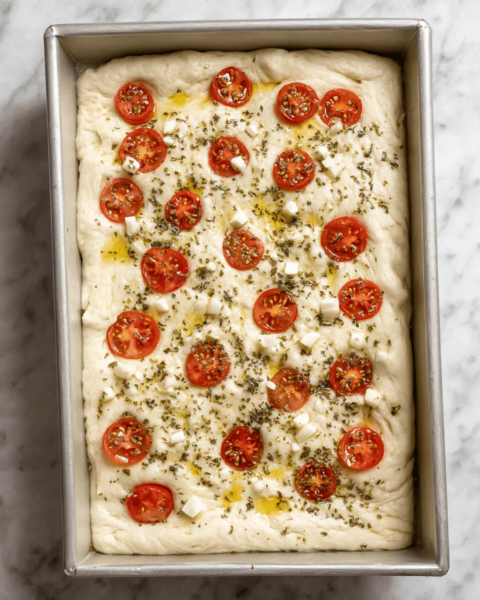 A rectangular baking pan filled with light beige dough spread evenly with a soft, bumpy texture. Small red cherry tomato slices are scattered on top along with small white chunks of garlic, and green dried herbs sprinkled all over. There are glossy drops of olive oil visible, giving a slight shine to the surface. The pan is placed on a white marbled texture surface. Photo taken with an iphone --ar 4:5 --v 7