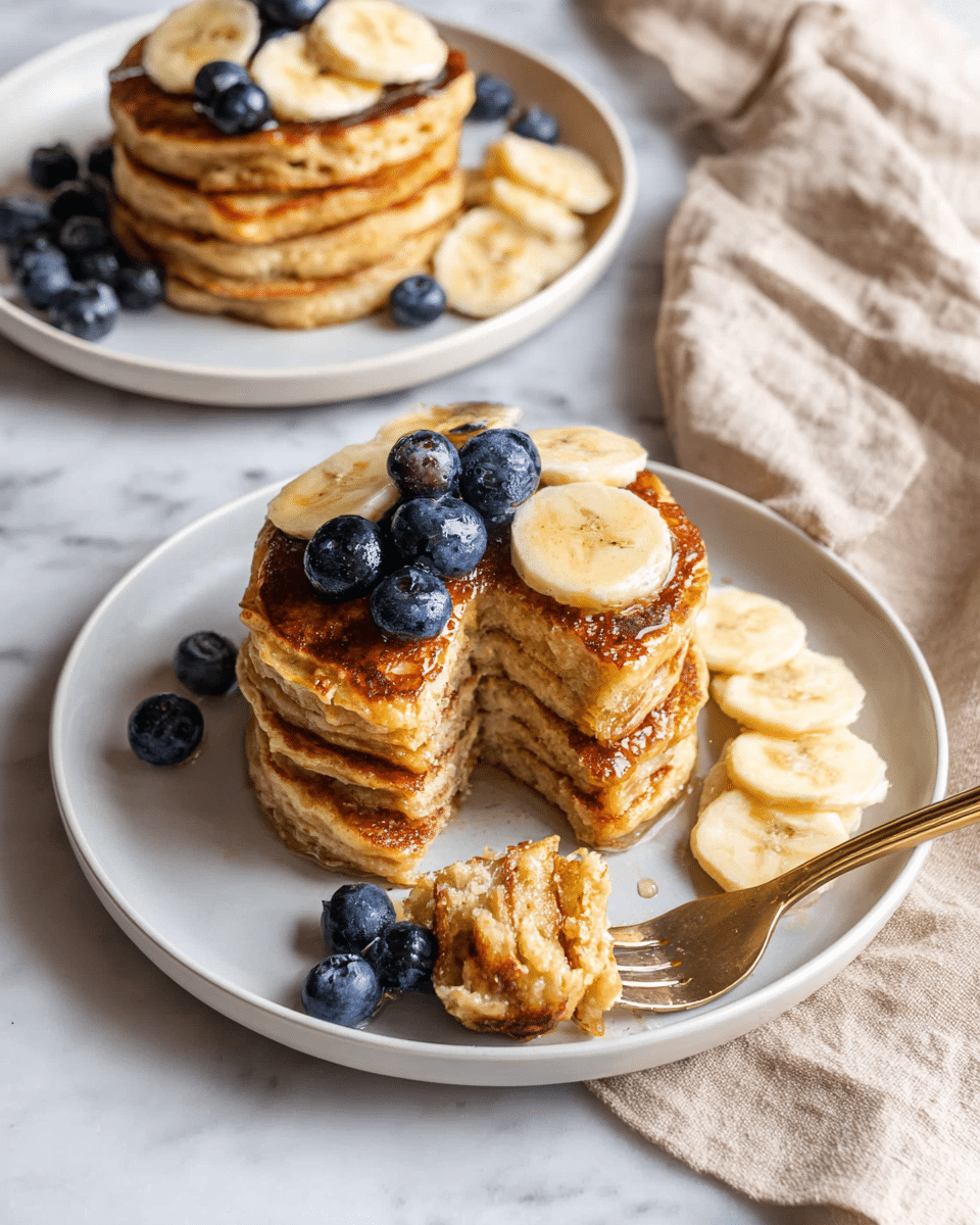 A stack of four golden-brown pancakes sits in the center of a white plate, with a slice taken out showing the thick, soft layers inside. The top pancake is decorated with three banana slices, each topped with shiny, plump blueberries. To the right of the stack, several banana slices are laid out on the plate. On the left side, a small pile of blueberries rests next to a fork holding a bite of pancakes, banana slice, and blueberry. In the background, another white plate holds a taller stack of pancakes with banana and blueberries on top, all placed on a white marbled surface with a beige cloth nearby. Photo taken with an iphone --ar 4:5 --v 7
