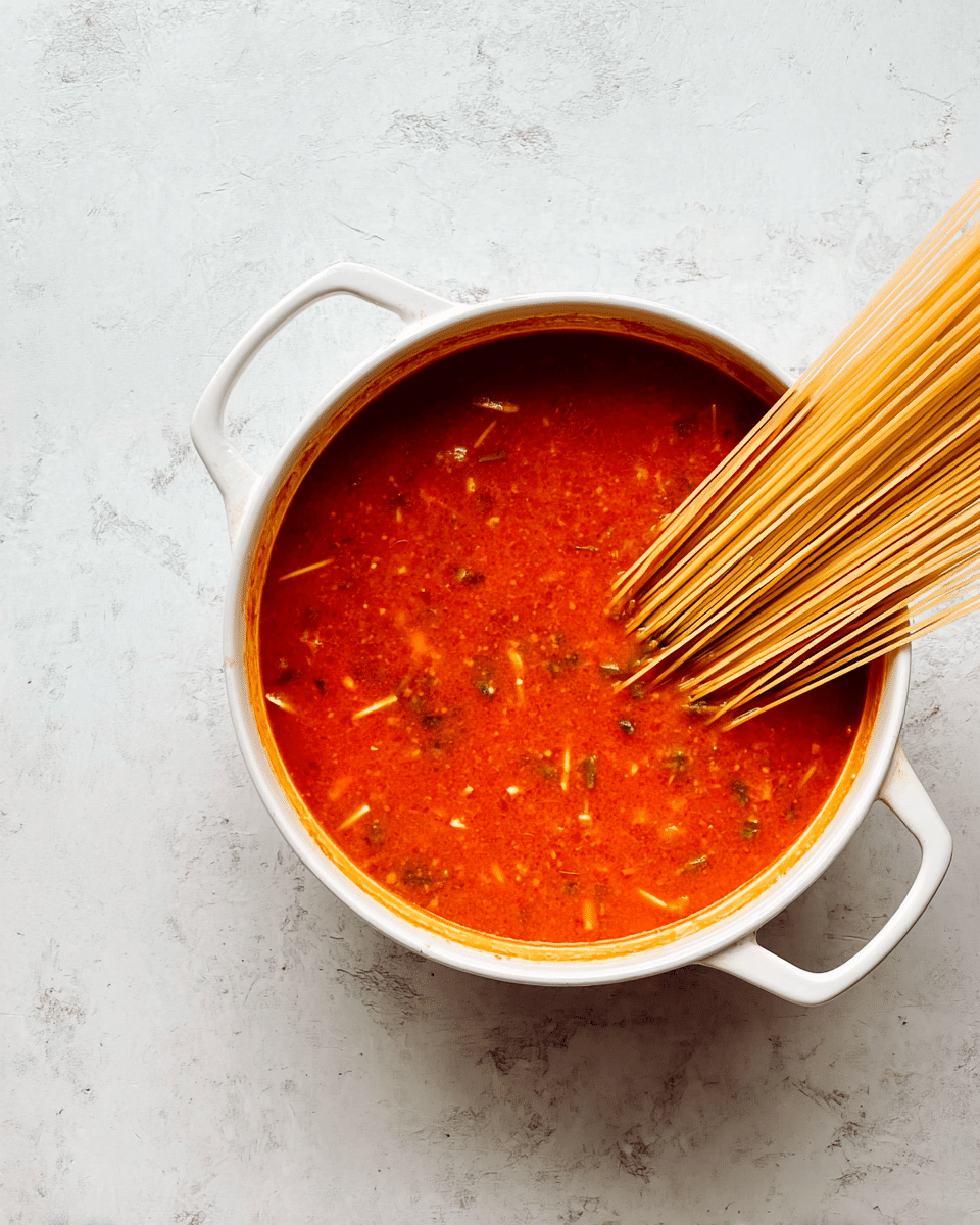 A white pot with two handles is filled with a thick red-orange sauce with small bits of herbs or spices visible throughout. Part of a bundle of dry pale yellow spaghetti noodles sticks out diagonally from the sauce, resting mostly on the right side with the ends partially submerged. The pot sits on a white marbled surface that has a slightly worn, rustic look. photo taken with an iphone --ar 4:5 --v 7