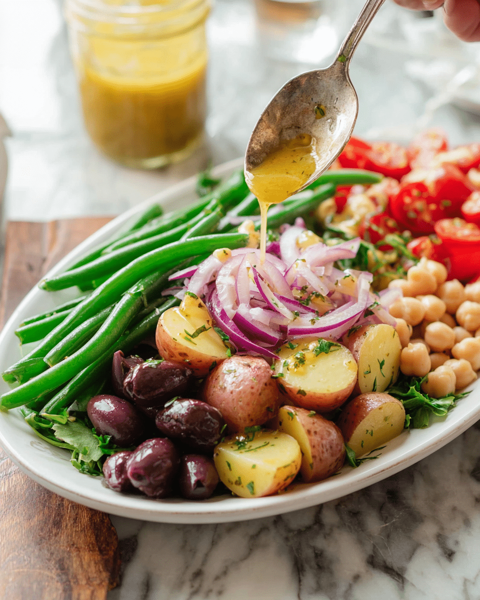 The dish is served in a white oval plate filled with several colorful layers. At the bottom, there is a green leafy layer, topped with green beans that are bright and fresh. On top of the green beans, there are small, halved red potatoes with a light yellow inside and specks of green herbs. Dark purple olives fill the front corner of the plate, adding a rich color contrast. To the side, thin slices of red onion are scattered, showing their light purple and white rings. Behind the onion, there are light beige chickpeas, and cherry tomatoes add bright red round shapes near the back. A woman's hand holds a spoon above the potatoes, drizzling a golden-yellow dressing over them. The plate sits on a white marbled surface with a jar of the same dressing in the background. photo taken with an iphone --ar 4:5 --v 7