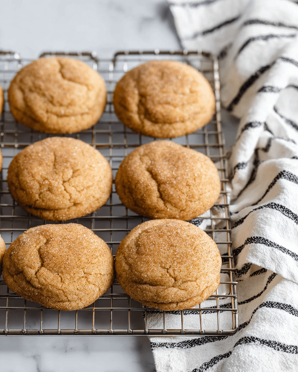 The image shows a group of round cookies with a golden brown color and a slightly cracked surface placed on a metal cooling rack. The cookies look soft and have a textured top with tiny sugar granules visible. The rack is set on a white marbled surface next to a white cloth with black stripes. The setting is simple and bright, focusing on the warm color and rough texture of the cookies photo taken with an iphone --ar 4:5 --v 7