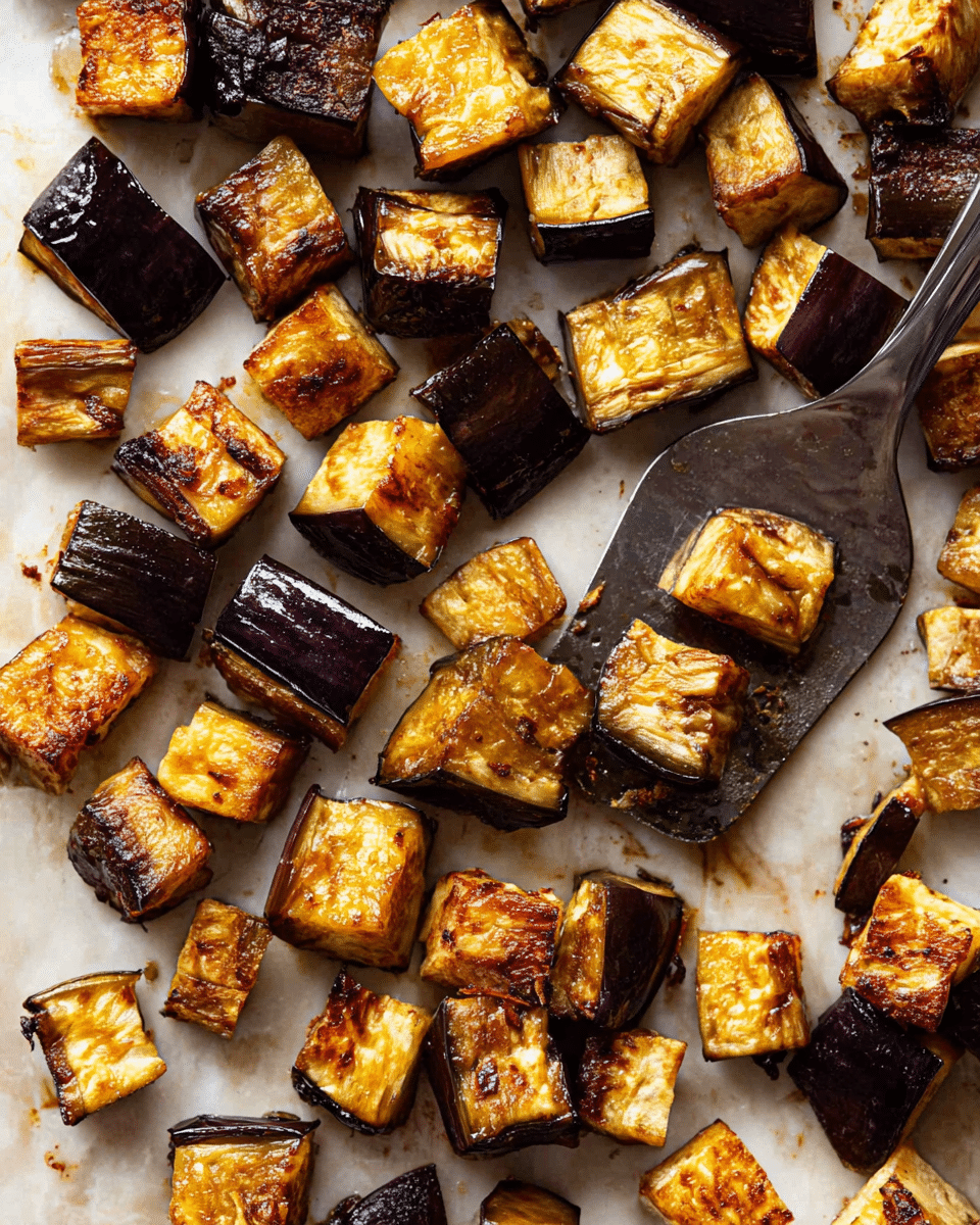 The image shows a white marbled surface covered with roasted eggplant cubes. Each cube is golden brown with slightly charred edges and a caramelized, tender inside. The eggplant skin is dark purple contrasting with the lighter, soft interior. A metal spatula is placed among the pieces, slightly lifting some cubes, showing their textured, cooked skin and a moist, soft center. The roasted eggplants are spread in a casual, natural way with some darker spots from caramelization. photo taken with an iphone --ar 4:5 --v 7