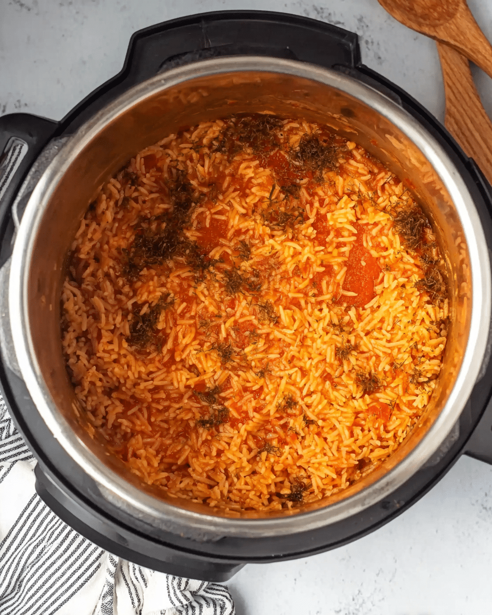 The image shows an inner pot of a pressure cooker filled with cooked rice mixed with tomato sauce, giving the rice an orange and red color. The rice looks soft and evenly spread inside the pot, with some darker bits of herbs or seasoning scattered around the edges. The pot is black on the outside, and it sits on a white marbled surface. Near the bottom right corner, there is a corner of a striped cloth and a wooden spoon visible. Photo taken with an iphone --ar 4:5 --v 7