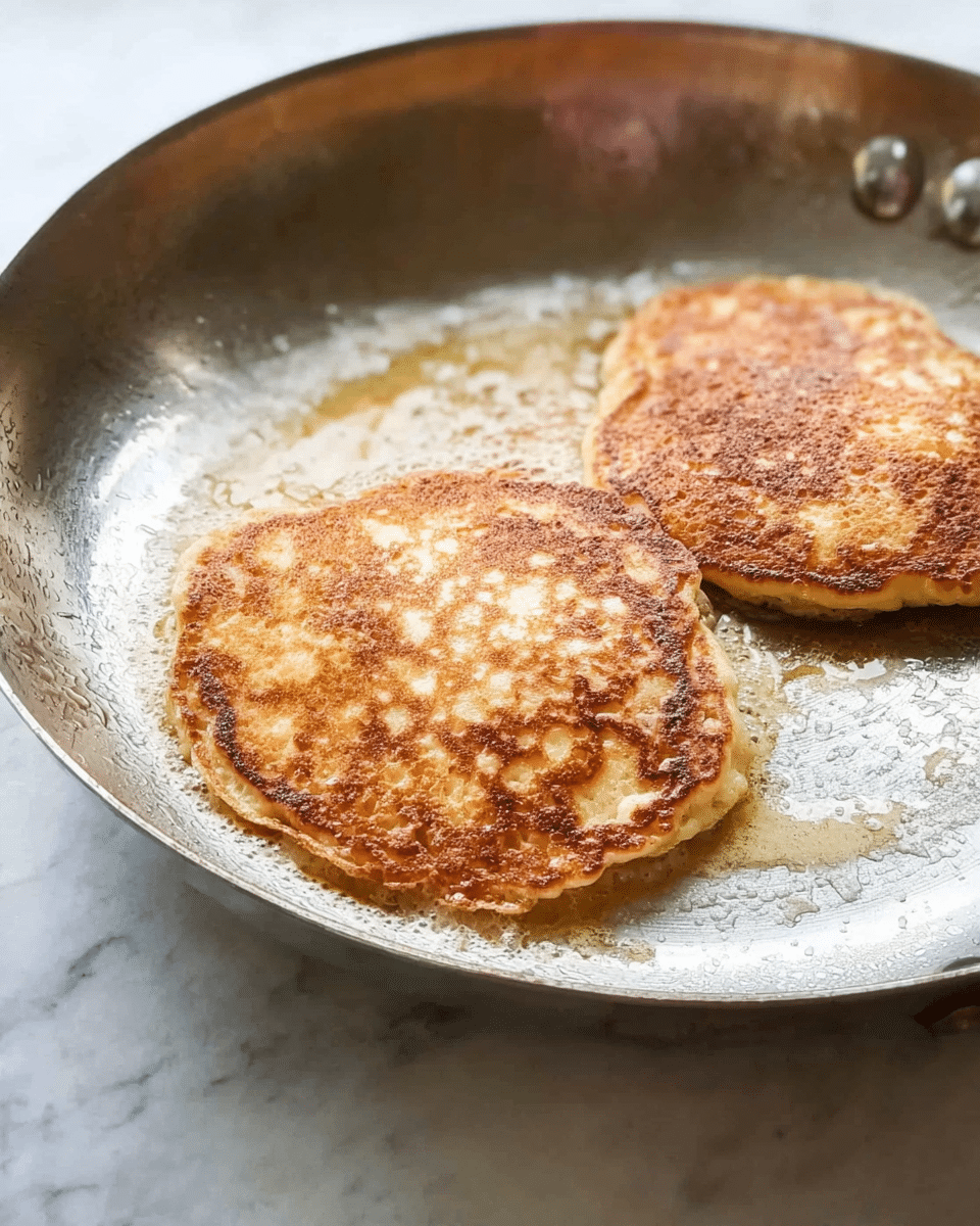 Two small pancakes are cooking in a silver frying pan. The pancakes are golden brown with a few darker spots, showing a soft and slightly rough texture. The edges are a bit uneven and the pan has some melted butter around the pancakes. The background is a white marbled texture. photo taken with an iphone --ar 4:5 --v 7