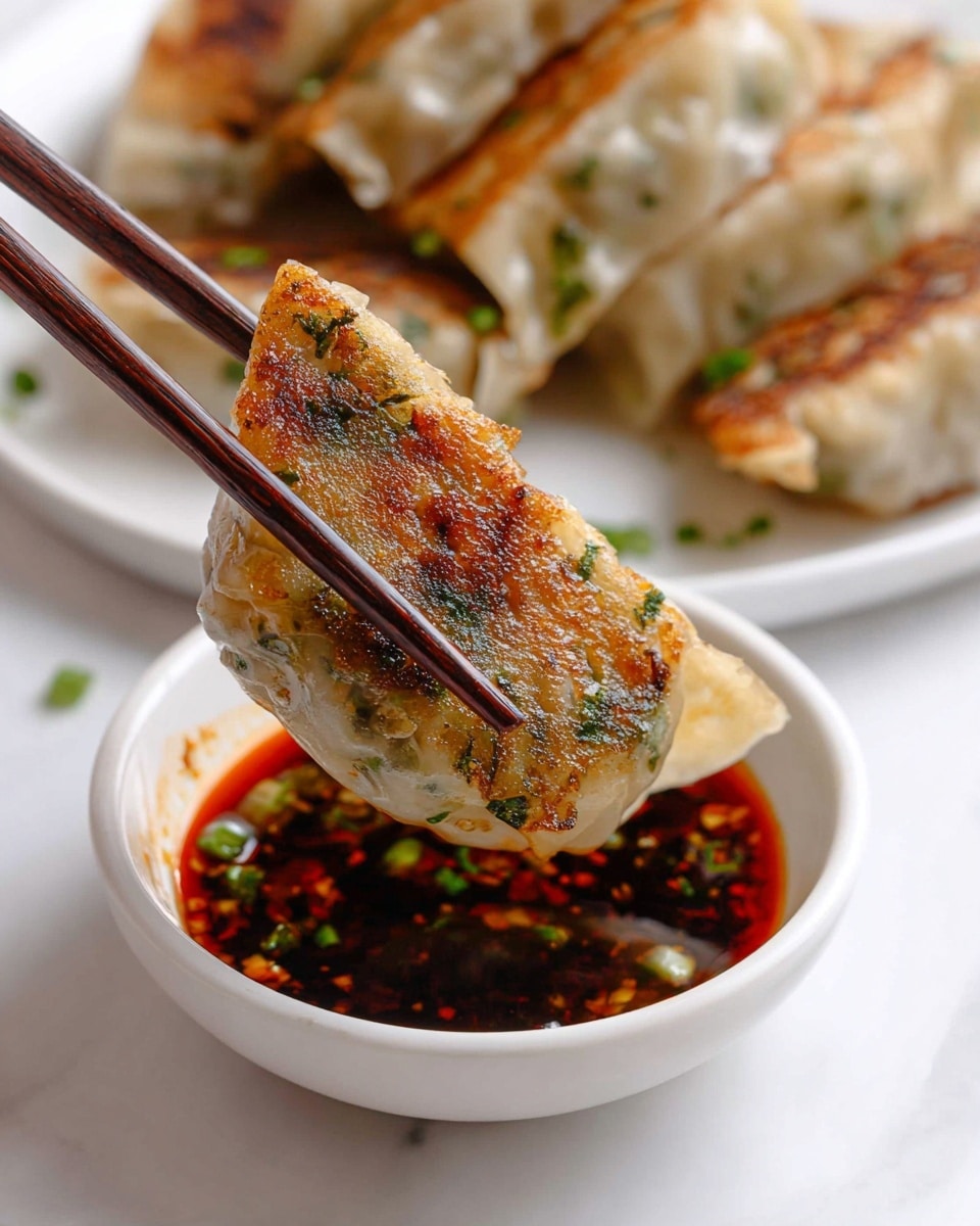 A grilled dumpling with a golden brown, slightly crispy bottom layer showing bits of green herbs. This dumpling has a translucent, soft white dough wrapper around the filling. The dumpling is held by dark brown chopsticks above a small white bowl filled with dark red chili oil sauce with scattered green chopped chives. In the background, more dumplings rest on a white plate, placed on a white marbled surface. Photo taken with an iphone --ar 4:5 --v 7