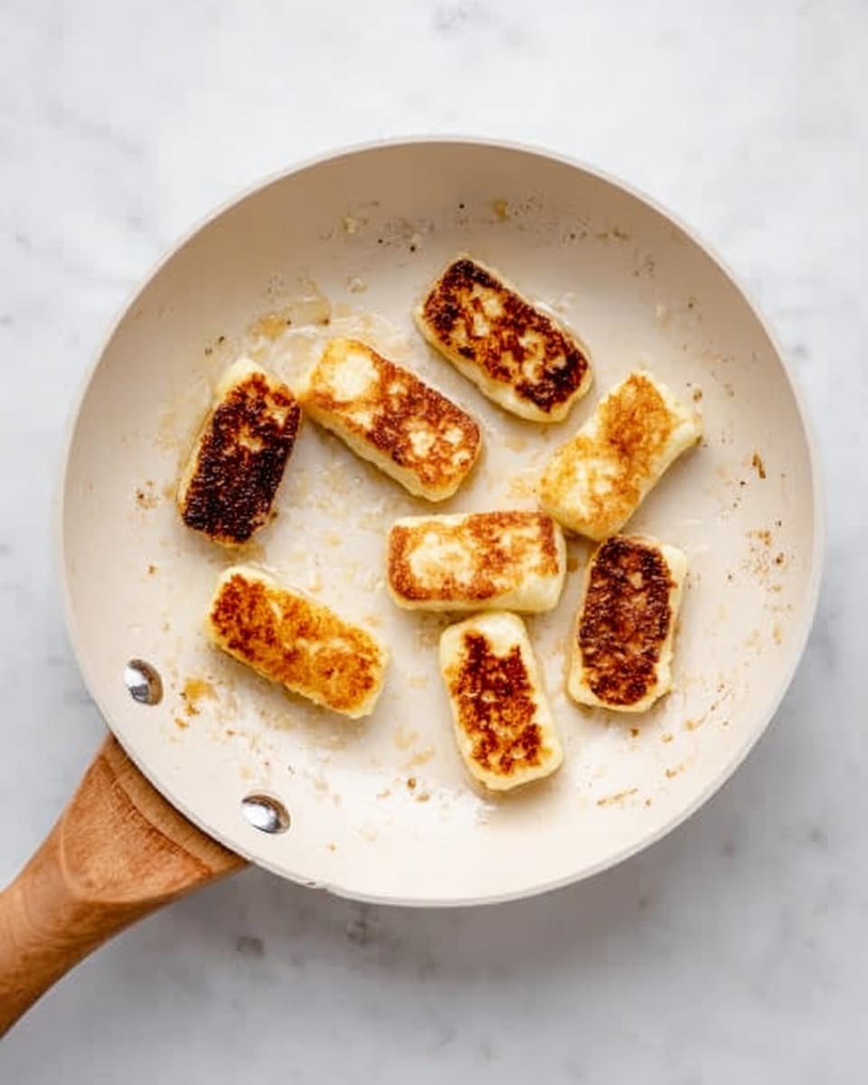 There is a white frying pan with a wooden handle placed on a white marbled surface. Inside the pan, there are nine small pieces of golden-brown fried cheese, each with a crispy, slightly darkened crust on one side and a light creamy color on the other. The pan has some brown spots from cooking around the cheese pieces, showing where the frying has taken place. The pieces are spread out unevenly across the pan. photo taken with an iphone --ar 4:5 --v 7