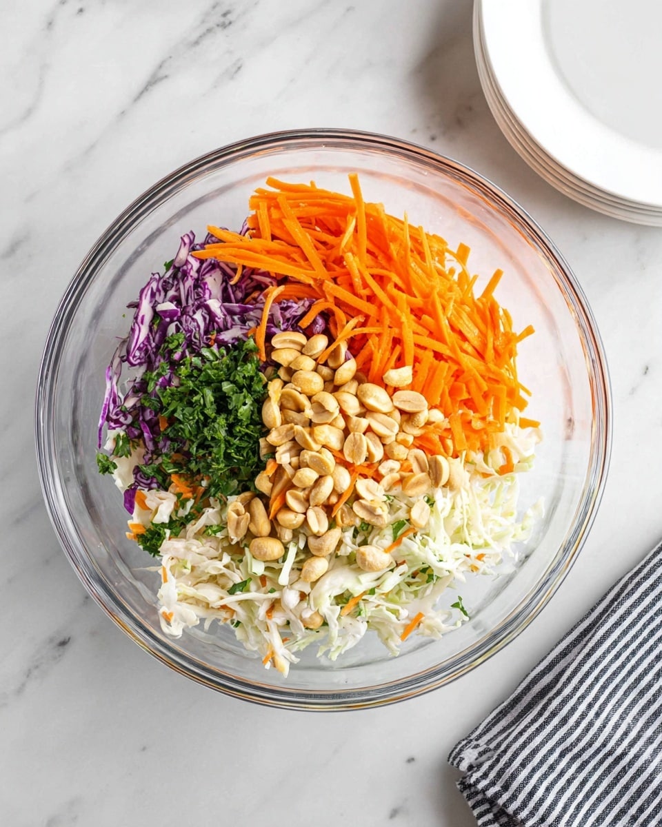 The image shows a clear glass bowl on a white marbled surface, filled with four main layers of ingredients arranged side by side rather than mixed. One layer contains thin orange carrot strips, another has chopped green herbs, a third layer shows whole light brown peanuts, and the last is a mix of shredded white and purple cabbage with some shredded orange carrots. A white plate is partially visible in the top right corner, and a striped black and white cloth is on the lower right side. photo taken with an iphone --ar 4:5 --v 7