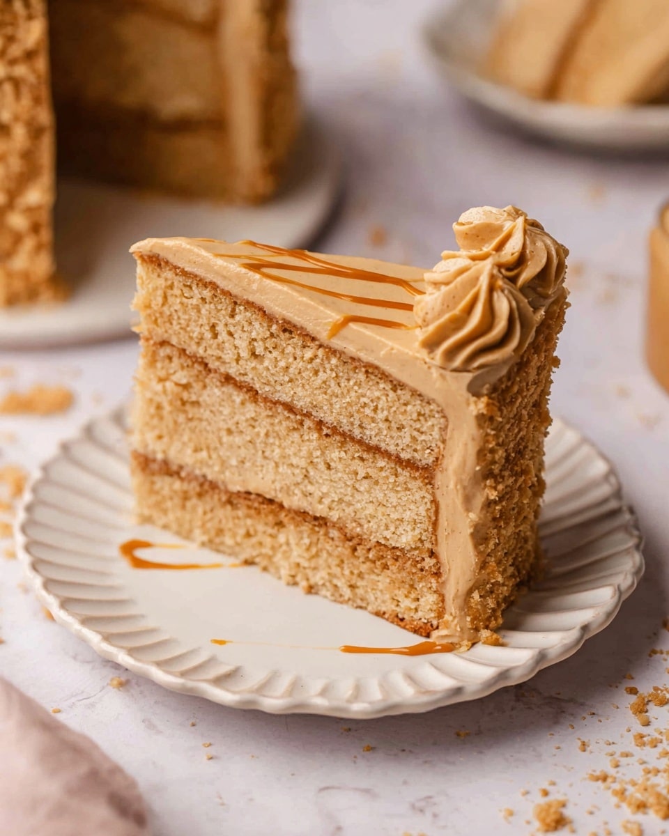 A slice of three-layer cake with light brown sponge and smooth, thick tan frosting between each layer is placed on a white scalloped ceramic plate. The top of the cake slice has a swirl of frosting with a small drizzle of caramel or butterscotch on it. The background is a white marbled surface with cake crumbs scattered around, and part of the full cake with a crumb border visible blurred in the background. photo taken with an iphone --ar 4:5 --v 7