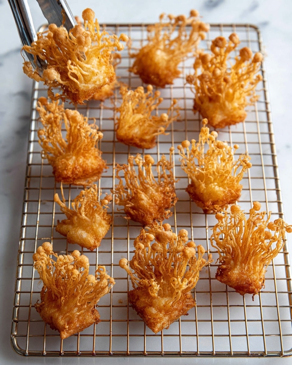The image shows ten pieces of deep-fried enoki mushrooms arranged loosely on a silver wire rack. Each piece has a golden-brown color with a crispy texture, featuring long, thin, branched stems with rounded tips, clustered tightly at the bottom base which is square-shaped and thicker. The wire rack is rectangular and placed on a white marbled surface, allowing the mushrooms to cool and stay crispy. One piece is held above the rack by a silver tong at the top left. The lighting highlights the crunchy texture and warm tones of the fried mushrooms. photo taken with an iphone --ar 4:5 --v 7