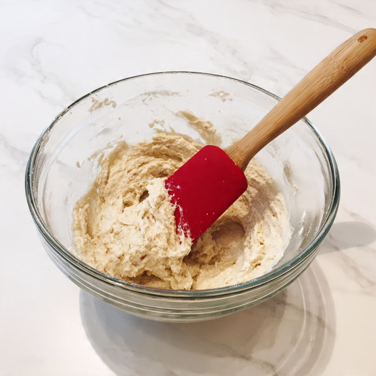 A clear glass mixing bowl filled with light beige, thick, and slightly lumpy batter sits on a white marbled texture. A red silicone spatula with a wooden handle is partially dipped into the batter on the right side of the bowl. The bowl has a wide rim and a spout on the left side. Photo taken with an iphone --ar 4:5 --v 7
