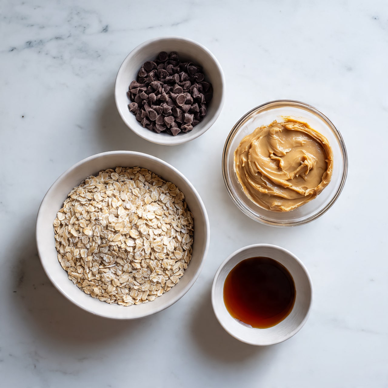 Four small white bowls with different ingredients sit on a white marbled surface. The largest bowl at the bottom left is filled fully with light beige rolled oats showing a dry, rough texture. Above and to the left is a smaller white bowl filled with dark brown, shiny chocolate chips that have a smooth texture. To the right of the chocolate chips is a clear glass bowl containing a creamy, light brown peanut butter with a smooth and thick texture. Below and to the right is another small white bowl with dark brown maple syrup, showing a shiny and slightly sticky surface. Photo taken with an iphone --ar 4:5 --v 7