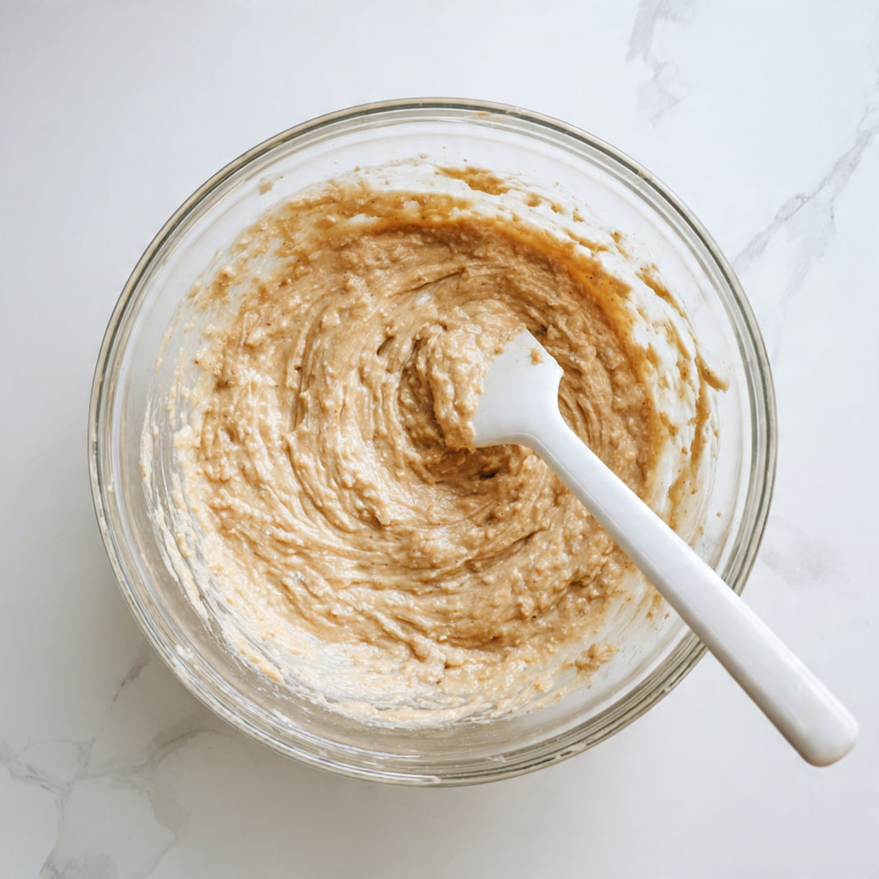 A clear glass bowl sits on a white marbled surface, filled with a thick, light brown batter that has a slightly rough texture with small lumps. A white spatula with batter clinging to it is partially submerged in the mixture, positioned inside the bowl on the right side. The batter has a swirled pattern, showing movement from stirring. In the top left corner, there is a white circle with the number 4 in bold black font. Photo taken with an iphone --ar 4:5 --v 7