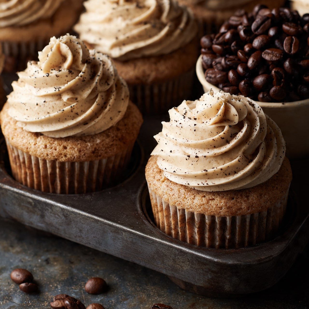 The image shows a close-up of four light brown cupcakes in a dark metal muffin tray with one cup filled with dark brown coffee beans. Each cupcake has one large swirl of creamy light brown frosting on top, textured with smooth ridges and specks of coffee powder sprinkled over it. The muffins have a slightly rough texture and are held in ribbed brown paper liners. The tray sits on a soft beige cloth on a white marbled surface. photo taken with an iphone --ar 4:5 --v 7