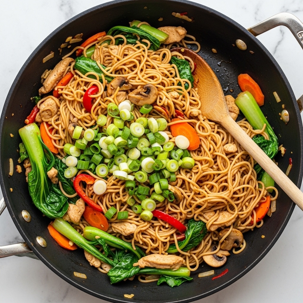 The image shows a large dark stir-fry pan filled with a colorful noodle dish. At the bottom, there are light brown thin noodles tangled together. Mixed in are bright green leafy vegetables and chopped light green spring onions scattered on top. Orange carrot slices and small red pepper strips add bright color. There are also light beige pieces of cooked chicken and small brown mushroom slices mixed in. The noodles and ingredients have a shiny texture from sauce. A wooden spoon is placed inside the pan, stirring the food. The pan sits on a white marbled surface. photo taken with an iphone --ar 4:5 --v 7