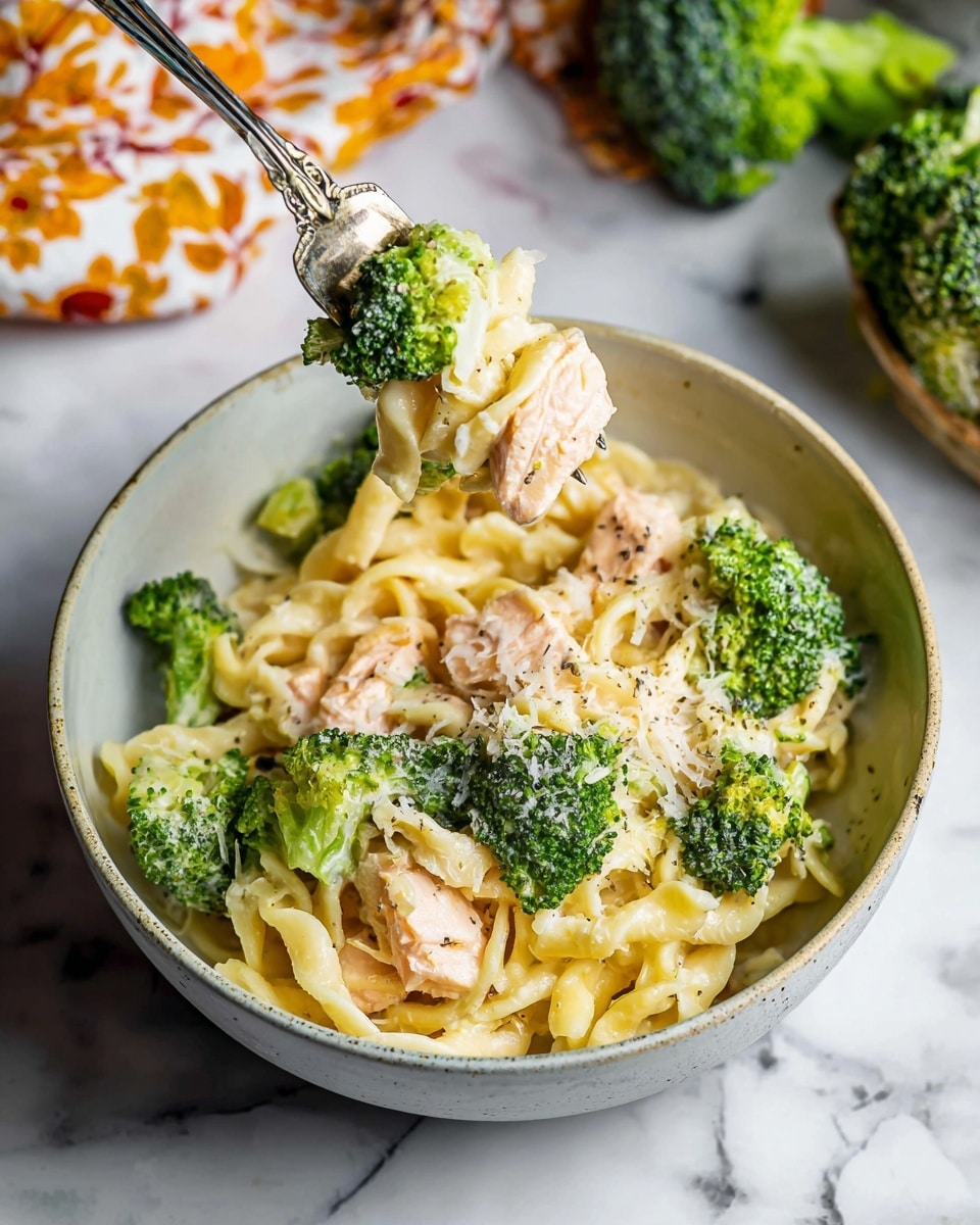 A white bowl filled with three main layers shows the dish: the bottom layer is creamy, light yellow pasta noodles twisted and coated with sauce, the middle layer has large, bright green broccoli florets scattered around, and the top layer features chunks of light pink cooked fish, all sprinkled lightly with grated white cheese and black pepper. A silver fork holds a piece of the fish and some noodles lifted above the bowl. The bowl rests on a white marbled surface with some broccoli and a cloth with an orange floral pattern nearby. photo taken with an iphone --ar 4:5 --v 7