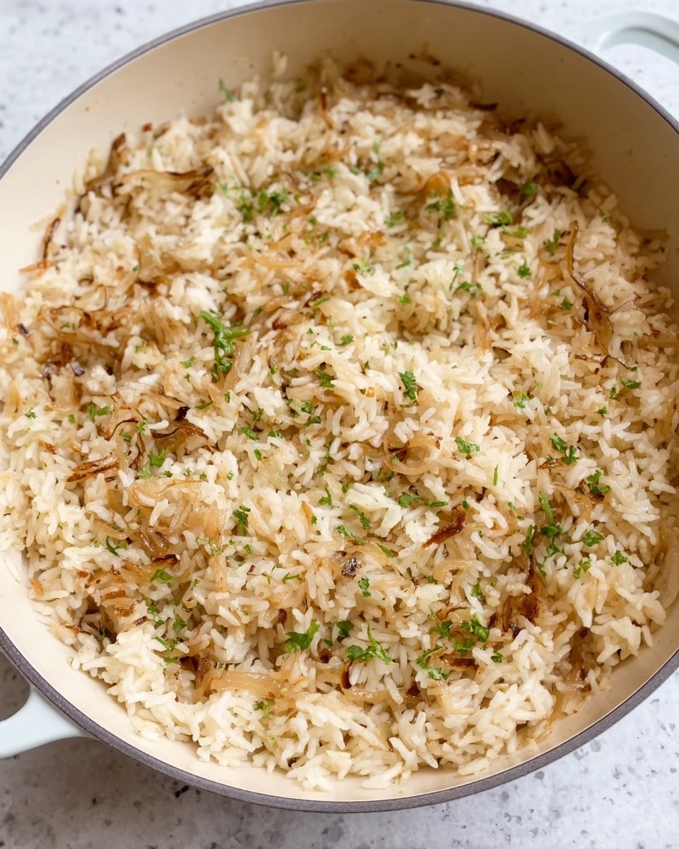 The image shows a close-up of cooked rice mixed with thin, soft caramelized onions spread evenly throughout. The rice is light beige with some brownish accents from the onions and has small green herb bits sprinkled on top for color contrast. The grains appear fluffy and separate, with no visible sauce or extra layers. This is all inside a light-colored pan placed on a white marbled surface. Photo taken with an iphone --ar 4:5 --v 7