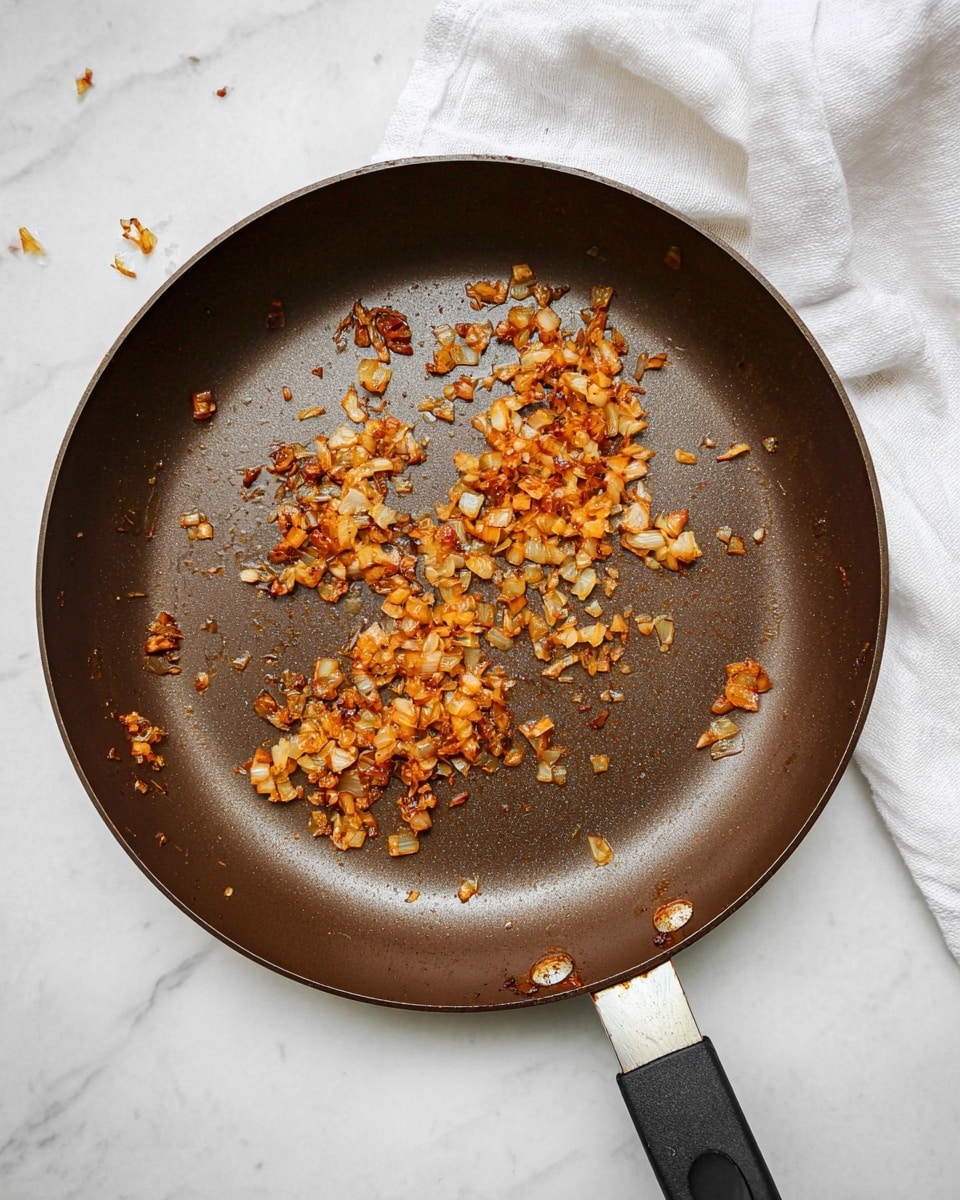 A dark brown frying pan with a black handle is placed on a white marbled surface, containing a single layer of small, chopped pieces of cooked onions spread unevenly across the middle. The onions are golden brown with some darker, caramelized spots showing a slightly crispy texture. There is a white cloth partially visible in the top right corner of the image. Photo taken with an iphone --ar 4:5 --v 7