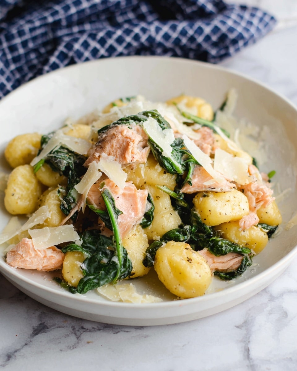 The dish shows a white plate filled with three main layers: soft yellow gnocchi as the bottom layer, topped with pieces of light pink cooked salmon and fresh dark green spinach leaves scattered throughout. Thin, pale shavings of cheese are spread on top, adding a delicate texture. The dish sits on a white marbled surface with a blurred blue checkered cloth in the background. The lighting is soft, making the colors look natural and warm. photo taken with an iphone --ar 4:5 --v 7