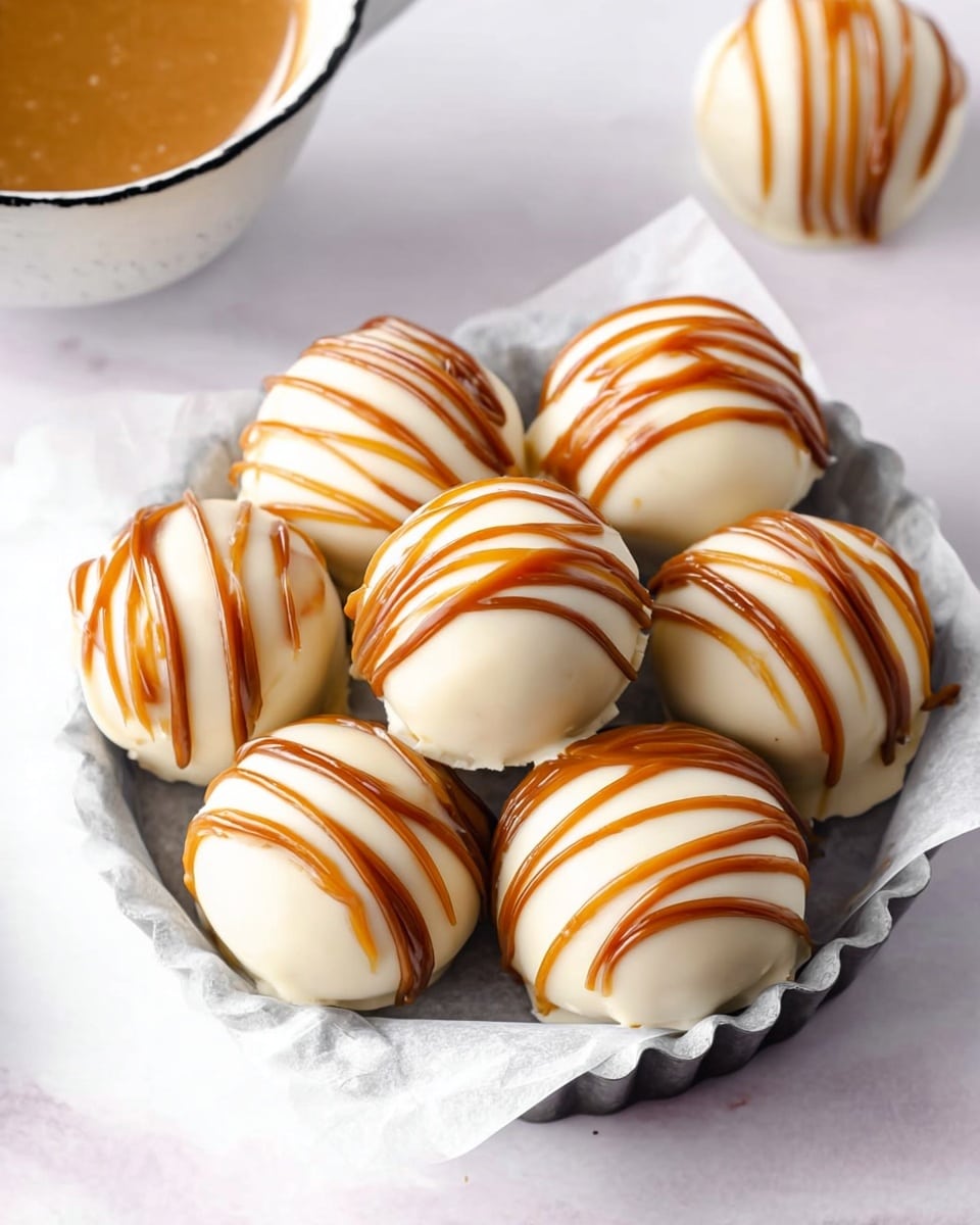 There are seven round white balls covered in smooth white chocolate with brown caramel drizzle lines on top, placed closely together on a sheet of white parchment paper inside a small metal fluted tart pan, which rests on a white marbled surface. Part of a cup filled with light brown caramel or sauce is visible on the top left, and a small white round ball with caramel drizzle is seen in the top right corner. The overall light setting highlights the creamy textures and rich caramel contrast. photo taken with an iphone --ar 4:5 --v 7