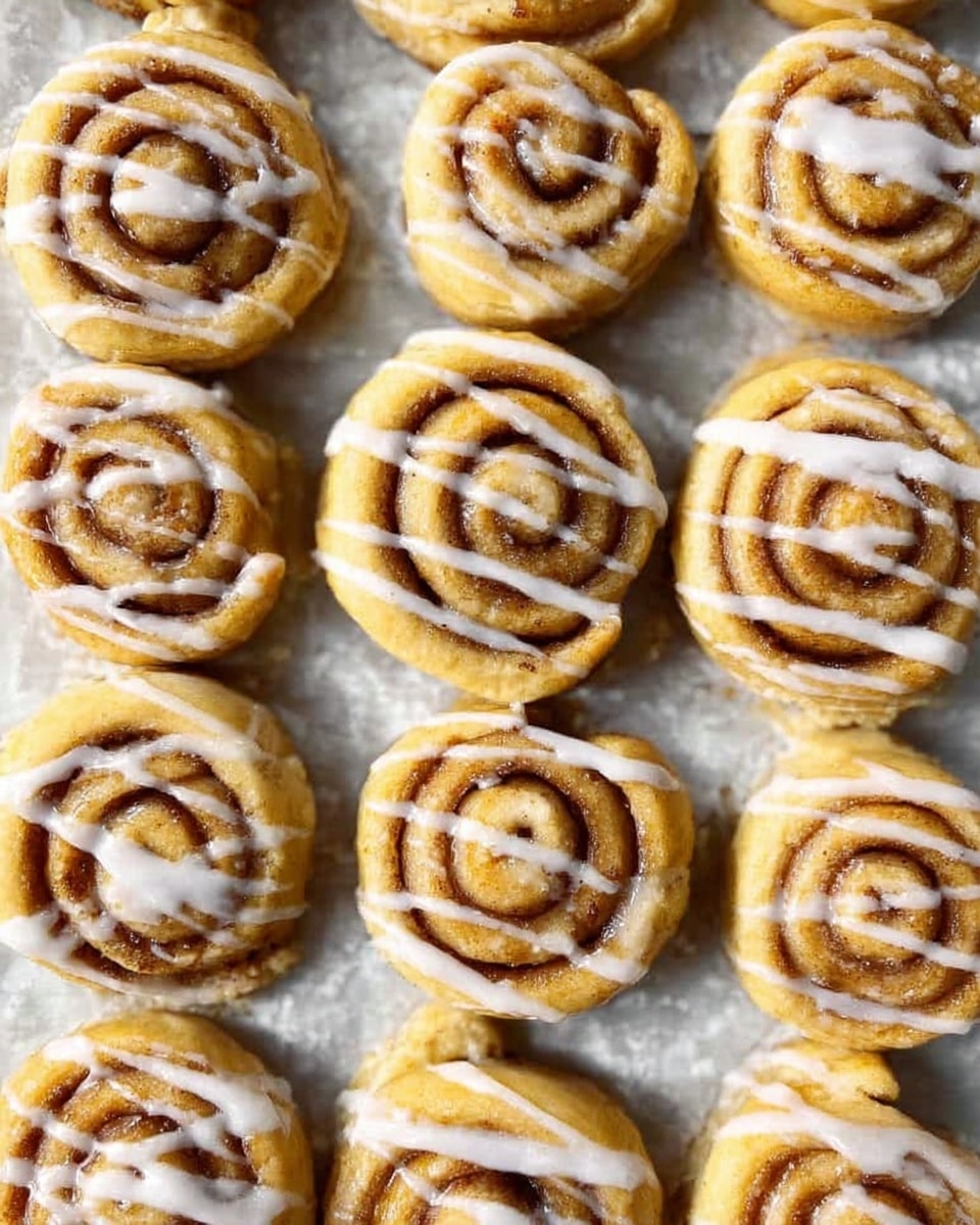 A close-up view of many small cinnamon rolls arranged in neat rows on a white marbled surface lined with parchment paper; each roll has visible spiral layers of golden-brown dough with cinnamon filling, topped with thin white icing lines drizzled across the tops in parallel patterns, creating a uniform look. photo taken with an iphone --ar 4:5 --v 7