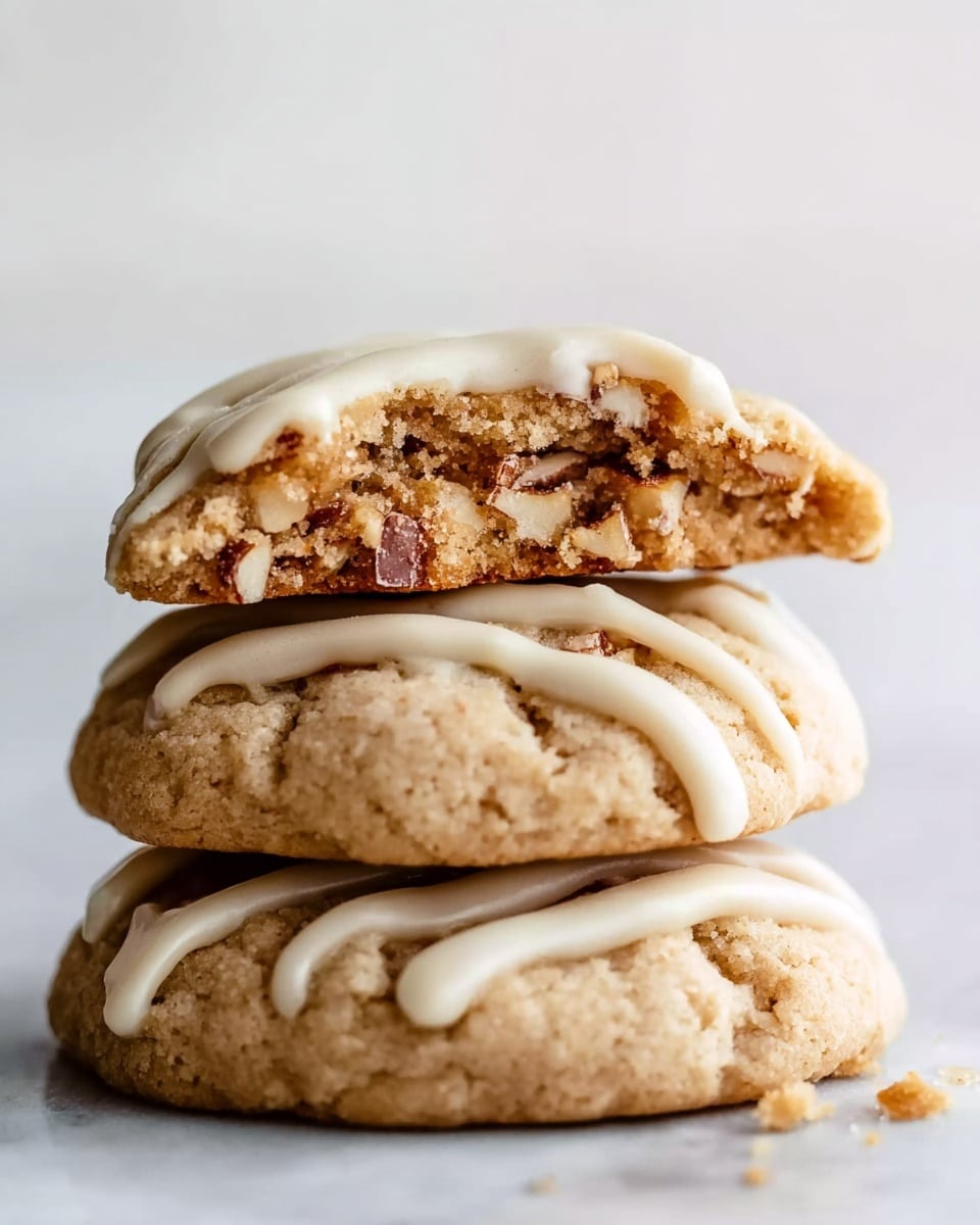 A stack of three light brown cookies with a soft texture is shown on a white marbled surface. The bottom two cookies have smooth white icing drizzled in thick lines across their tops. The top cookie is slightly broken, revealing a chewy inside with visible pieces of nuts or chocolate. The cookies look fresh and soft, with some crumbs around the base. Photo taken with an iphone --ar 4:5 --v 7