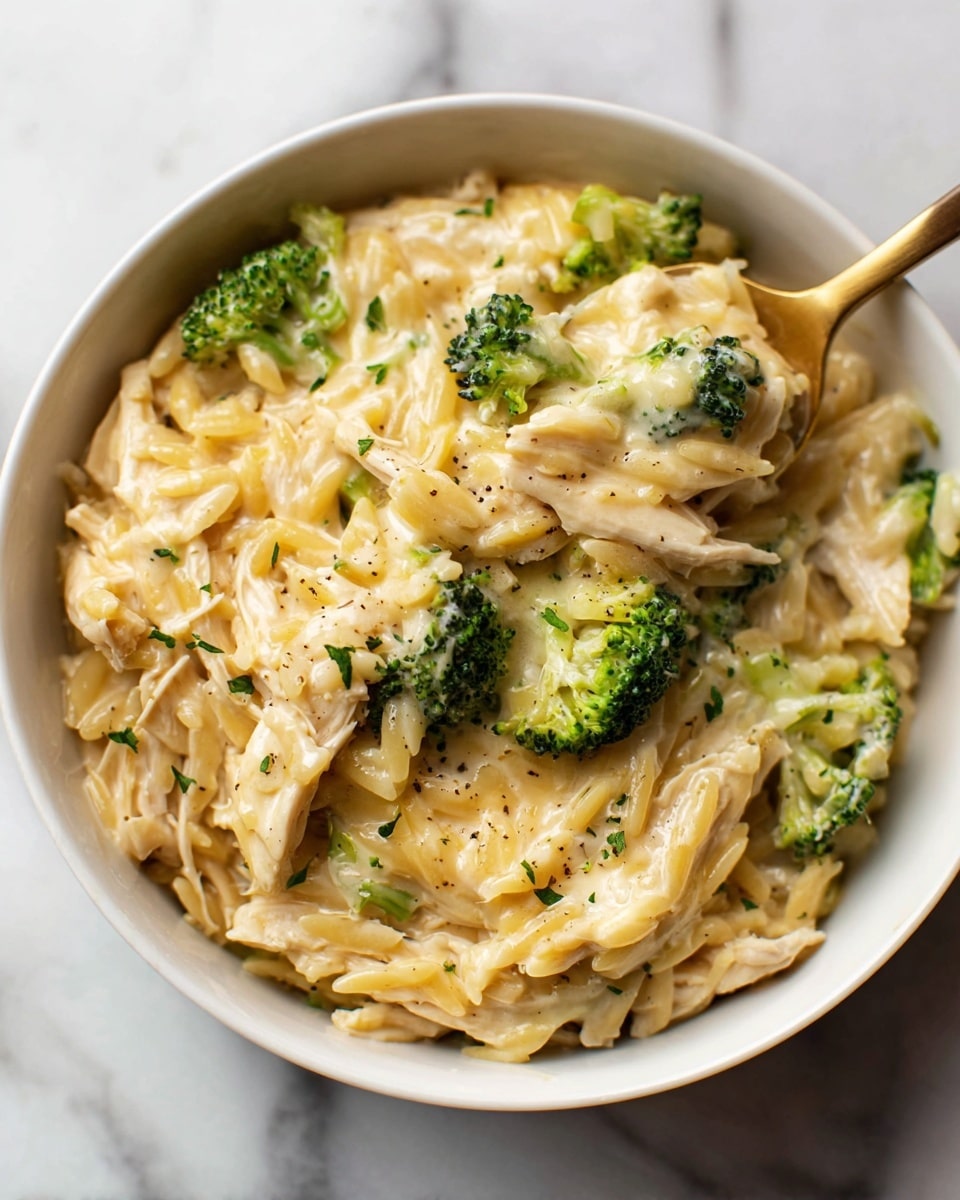 The image shows a white bowl filled with a creamy dish that has three main layers: shredded light beige chicken pieces, small bright green broccoli florets, and creamy, pale yellow orzo pasta mixed together. The sauce looks smooth and thick, coating all the ingredients evenly with a slight shine. The bowl is resting on a white marbled surface, and a gold and white spoon is partially dipped into the bowl, lifting some of the creamy orzo, chicken, and broccoli. The colors are warm and fresh, with visible bits of black pepper and small green herbs scattered on top. Photo taken with an iphone --ar 4:5 --v 7