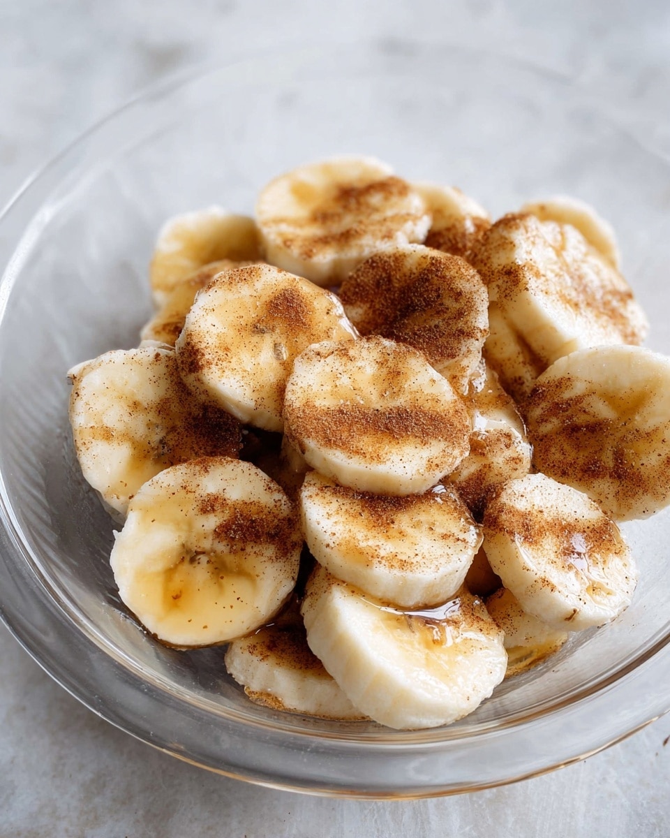 This image shows a close-up of a clear glass bowl filled with evenly sliced banana pieces. The bananas are creamy white with soft edges, and they are covered with a light layer of brown cinnamon powder and some liquid, likely honey or syrup, giving a shiny texture to parts of the banana slices. The bowl sits on a white marbled surface that adds a clean, soft background to the warm colors of the bananas and cinnamon. photo taken with an iphone --ar 4:5 --v 7